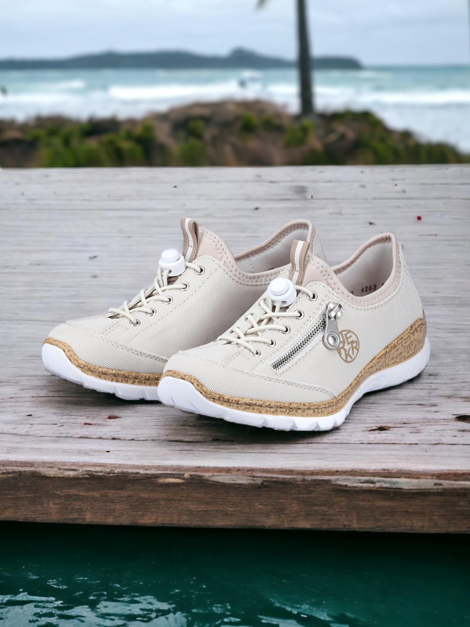 06802 Rieker Summer Days Sneaker Beige fra Rieker Comfort & Style har beige lerret, hvite såler, glidelåsdetaljer og memory-soft såler, sett utendørs på tre med et uklart hav og palmer i bakgrunnen.