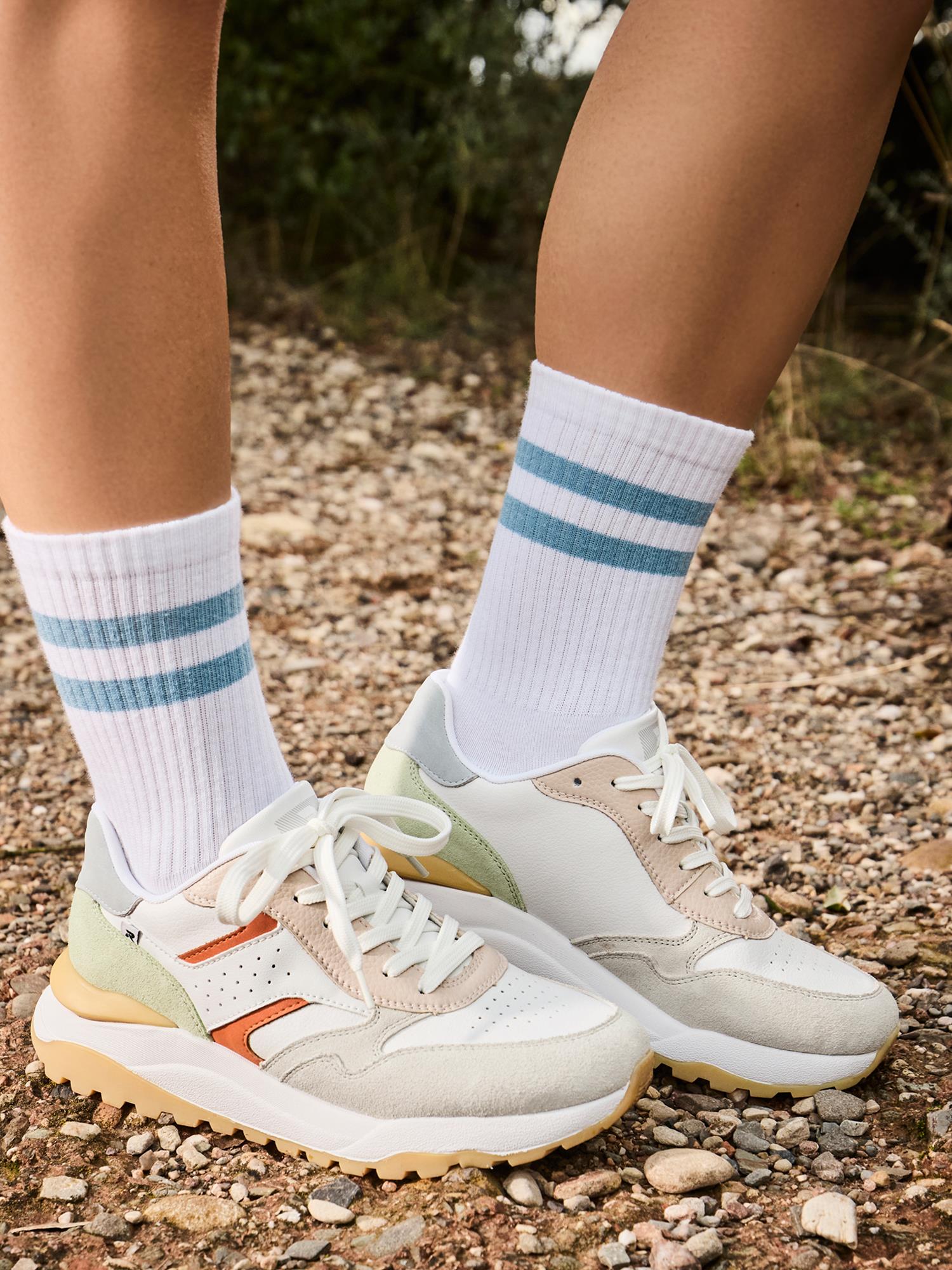 Utendørs på grus viser en person bare frem leggen og føttene i Rieker Sport 03546 TrailMotion Hvit Semsket Sneakers med pastellfargede detaljer og hvite sokker med to lyseblå striper, som understreker behagelig komfort.