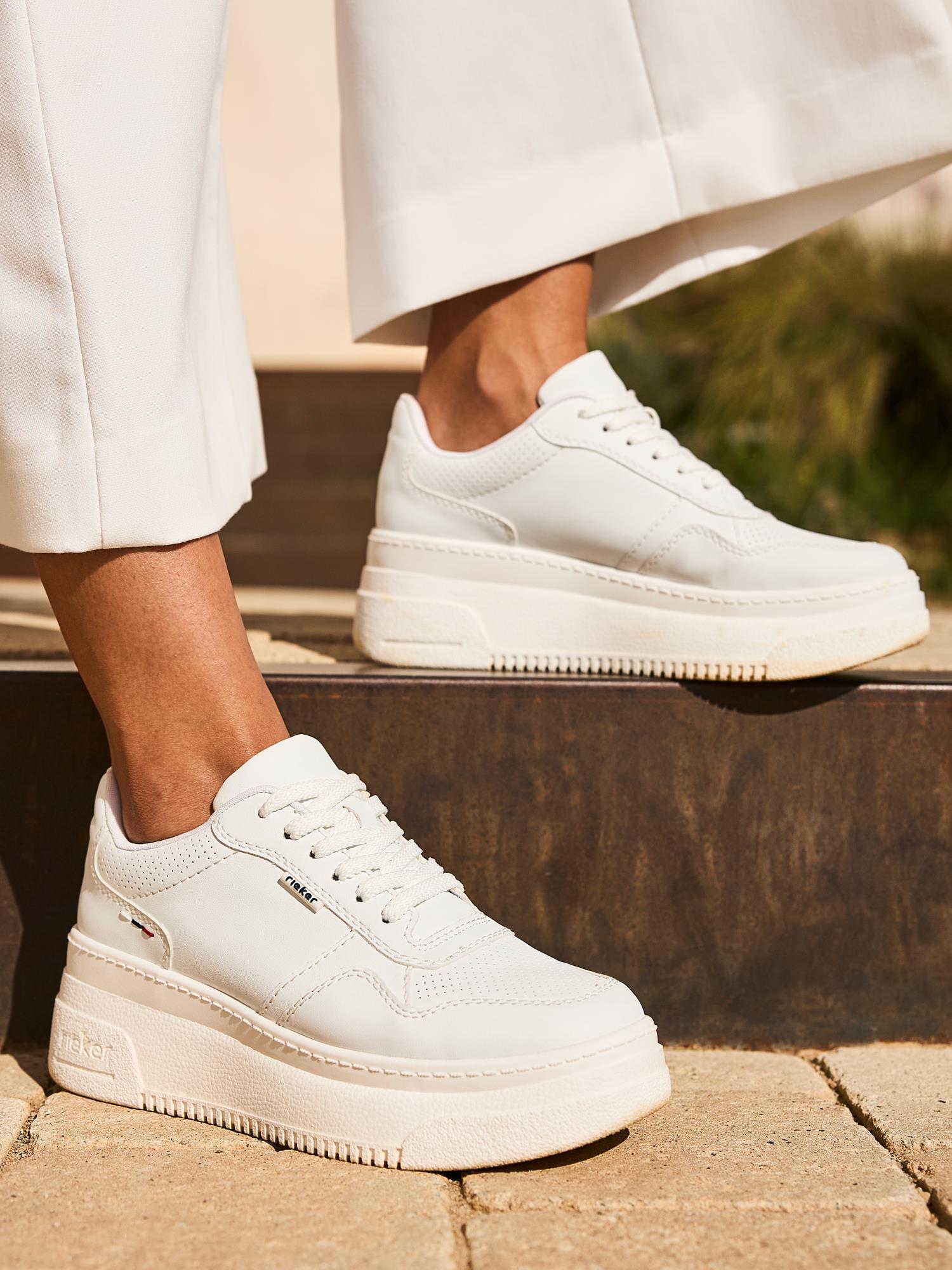 En person i hvite bukser med vide ben har på seg Rieker Comfort & Style 03280 Essential Comfort Sneakers med Antistress-teknologi, tykke teksturerte såler, hvite snørebånd og subtilt sidebranding mens hun står på en steintrapp utendørs.
