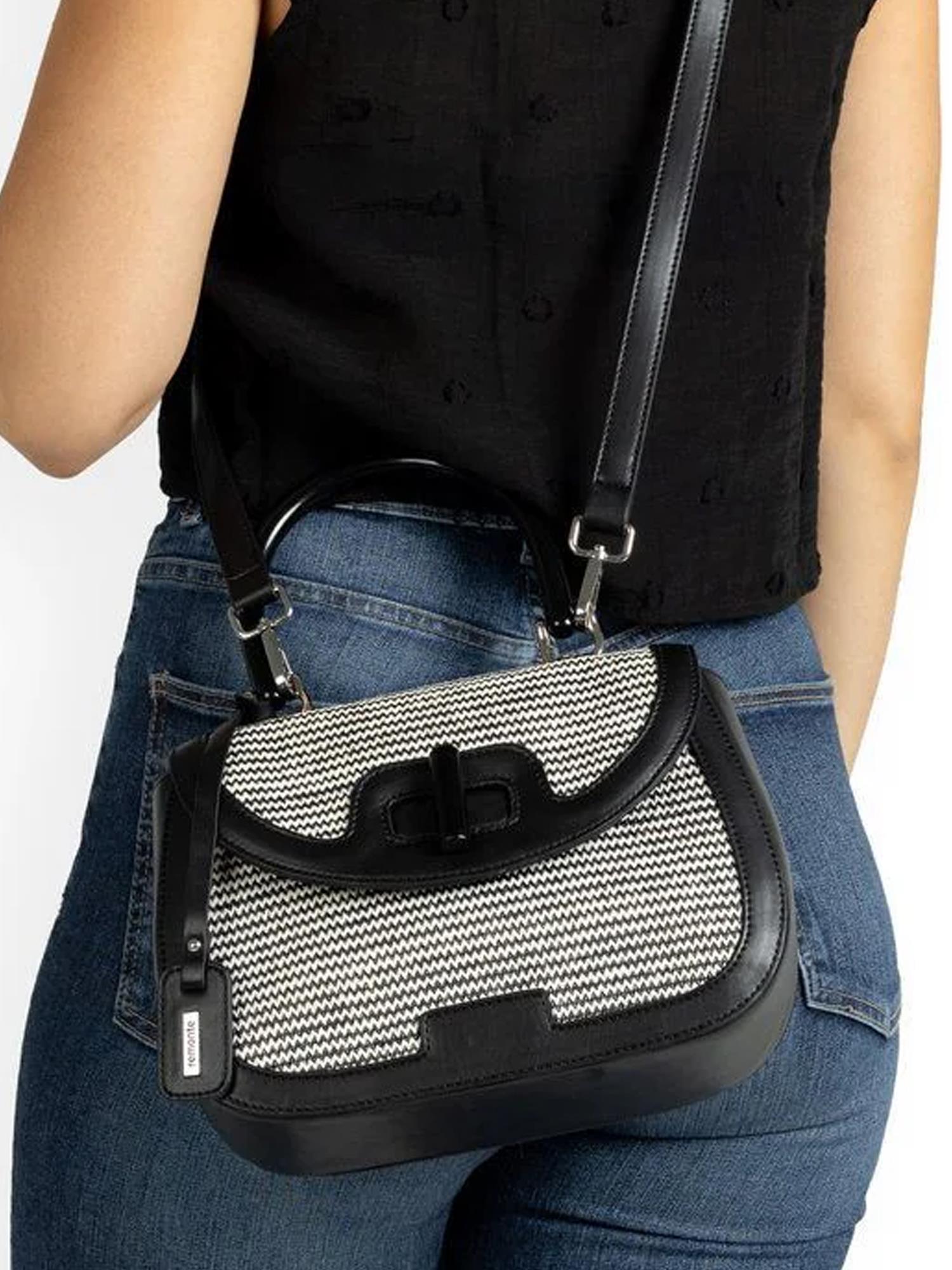 En person i blå jeans og svart ermeløs topp bærer en Remonte 03715 Rieker Siradia Handbag Svart med svart og hvitt mønster, kunstskinnrem og skinnhåndtak over skulderen.