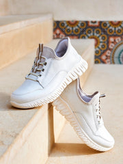 03263 Rieker White Ready2Go Slip-on fra Rieker Comfort & Style, med teksturerte såler og et komfortabelt slip-on-design, hviler på beige steintrapper med fargerike mosaikkfliser i bakgrunnen.