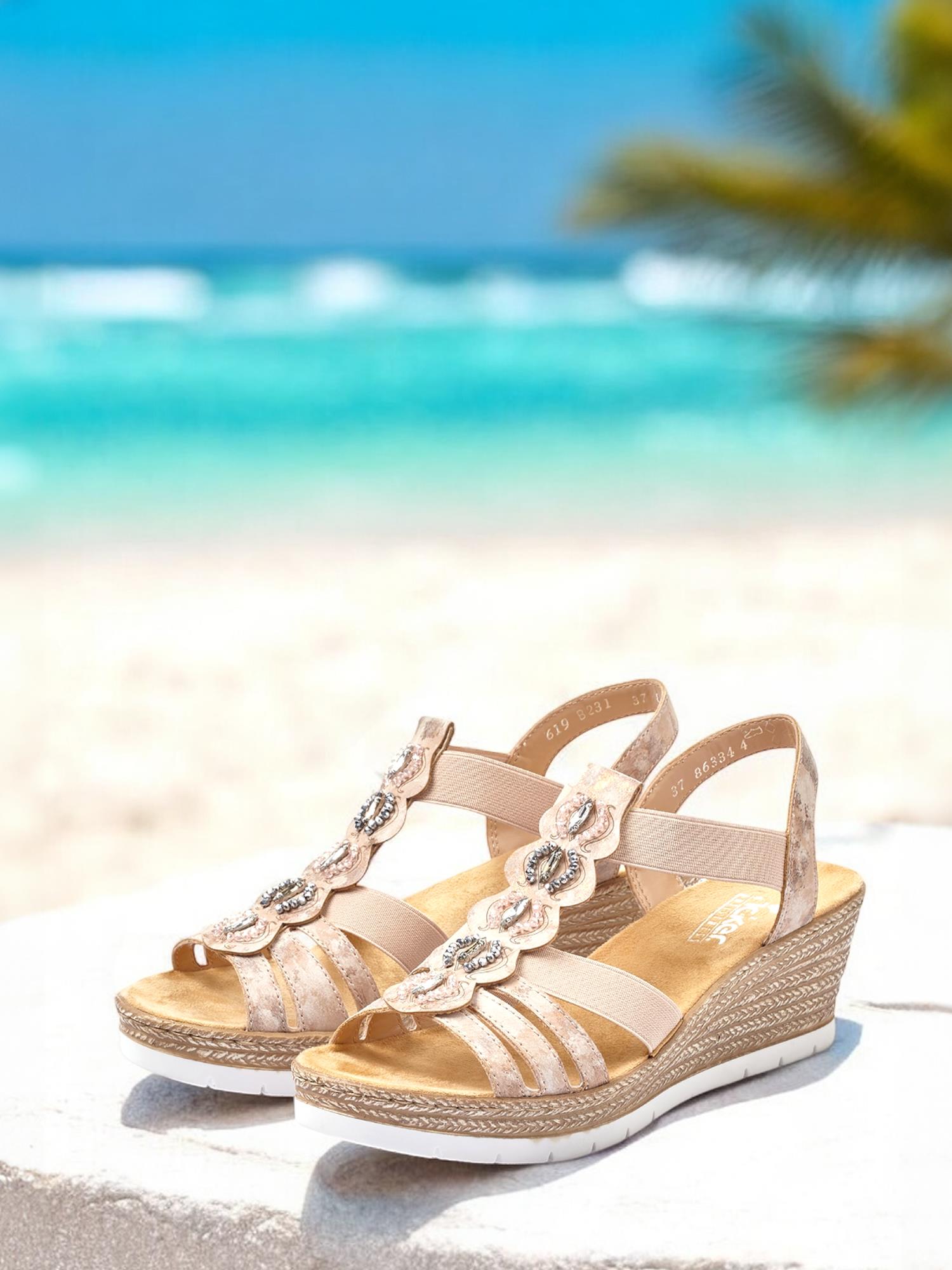 07636 Rieker Zahara Stone-sandalen fra Rieker Comfort & Style har en beige tone, høyere kilehæl og blomsterdetaljer, vist på hvitt med en uskarp strand, turkis hav og palme i bakgrunnen.