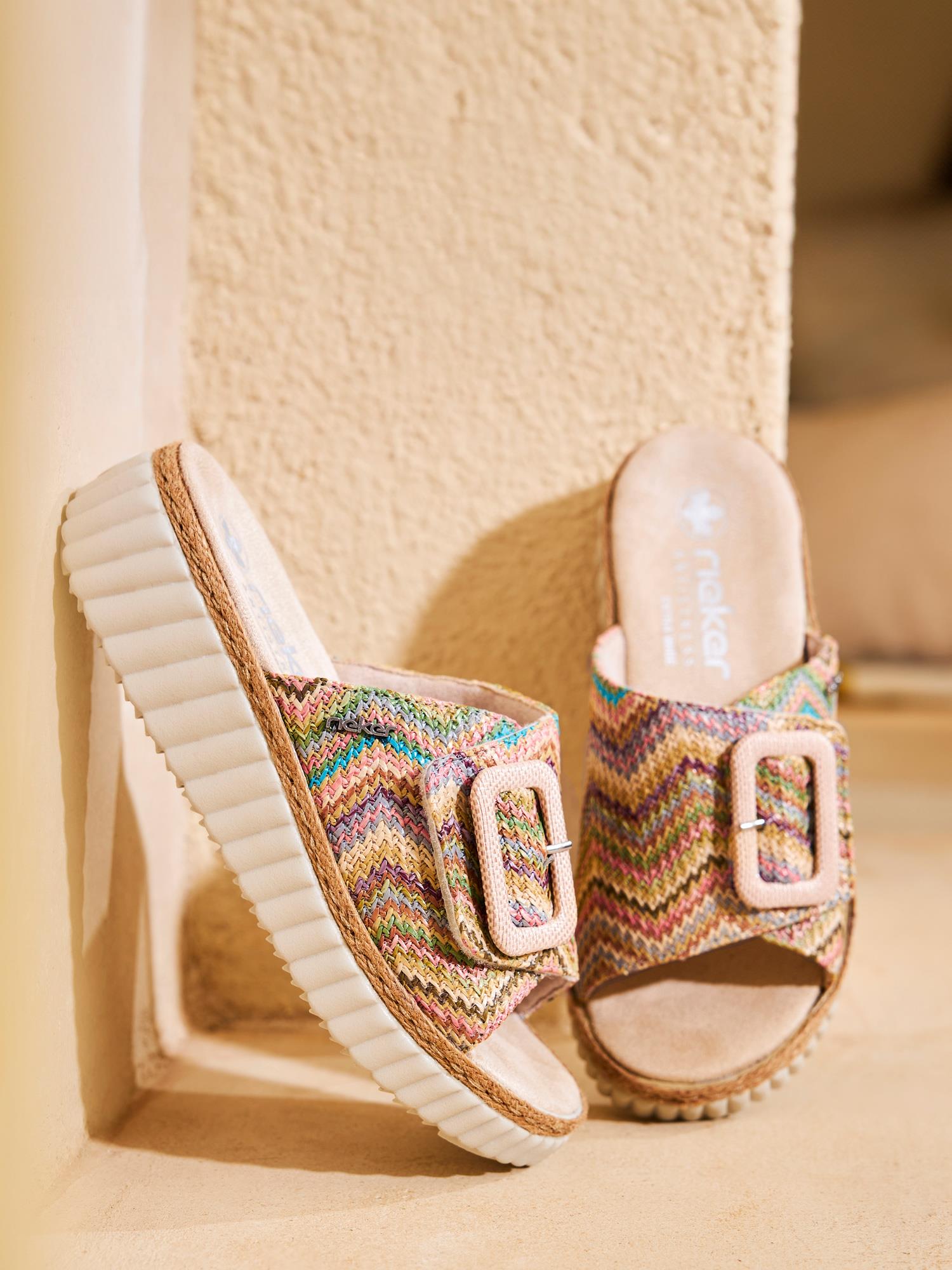 Mot en beige vegg står 03049 Rieker Solenya Slip-in Sandals fra Rieker Comfort & Style - fargerike, vevde slip-ins med store spenner og hvite, riflede såler, en oppreist og en skjev for å fremheve den ultralette komforten.