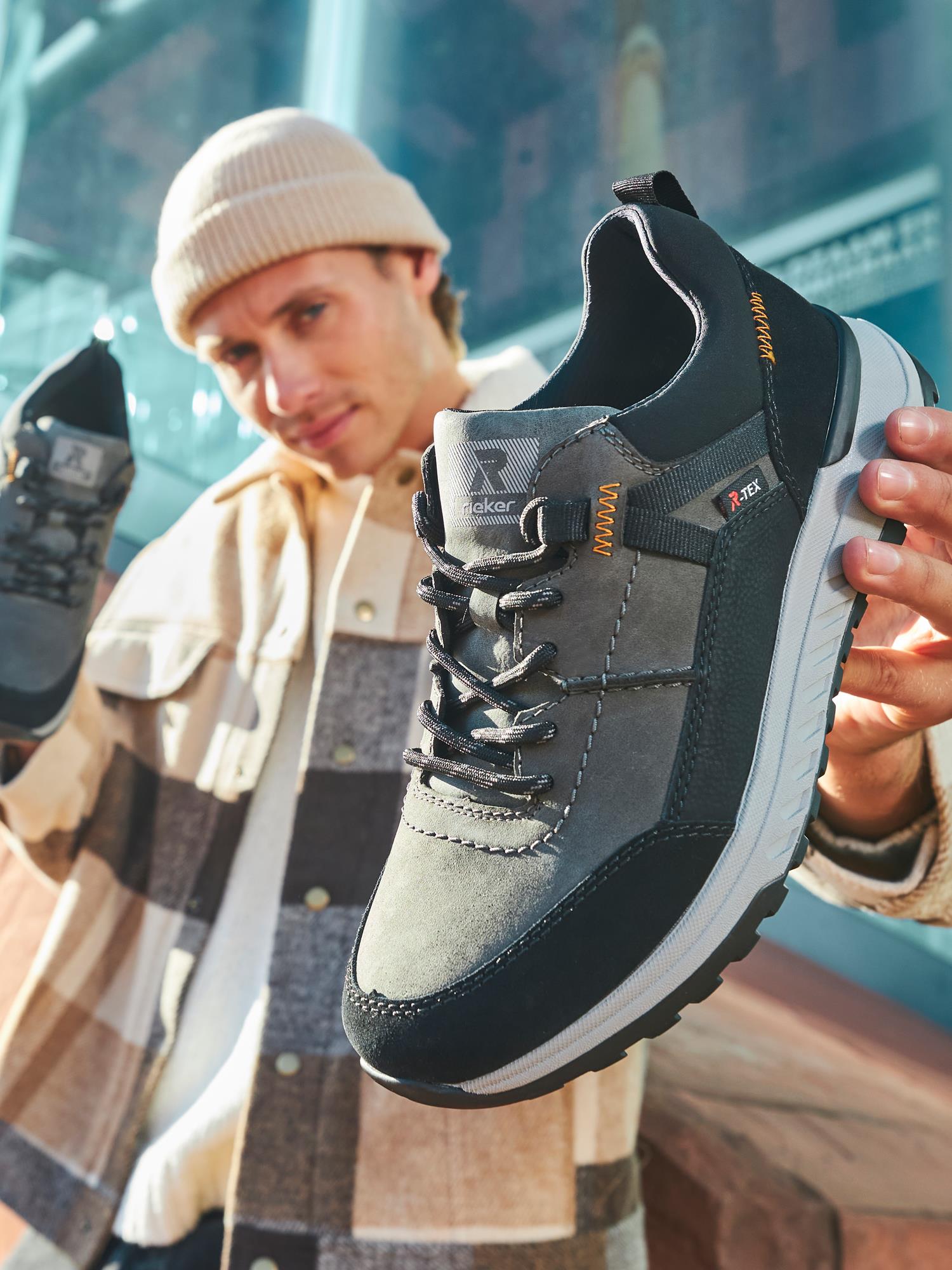 En mann i beige lue og rutete jakke holder en grå og svart Rieker Sport 04504 Rieker-Evolution Peak Sneaker Waterproof nær kameraet, og fremhever detaljene mot en uskarp urban bakgrunn.