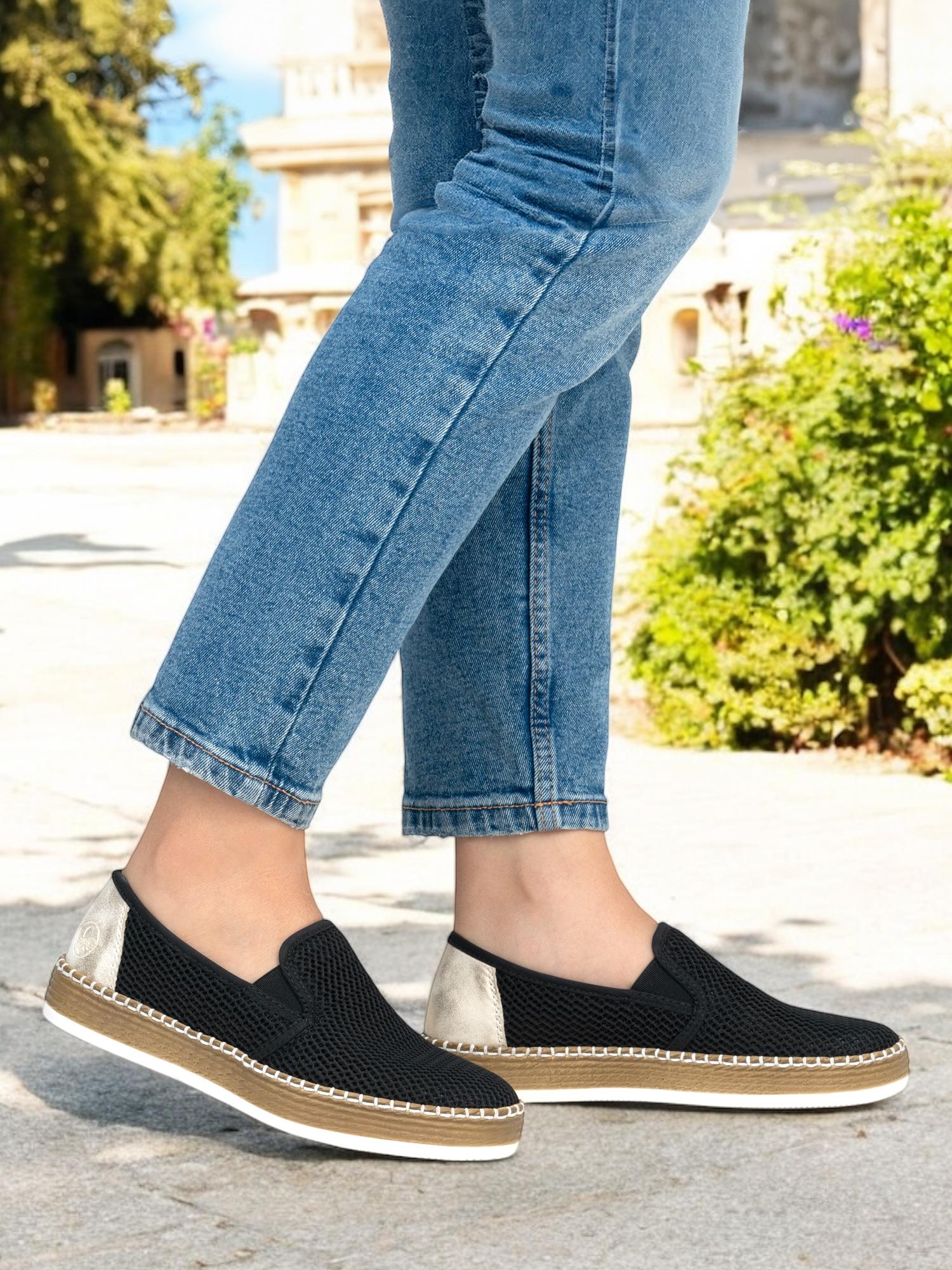 En person i blå jeans og 03156 Rieker BreezeWalk Sommersko by Rieker Comfort & Style står utendørs på en solrik dag, med grøntområder og en bygning i bakgrunnen.