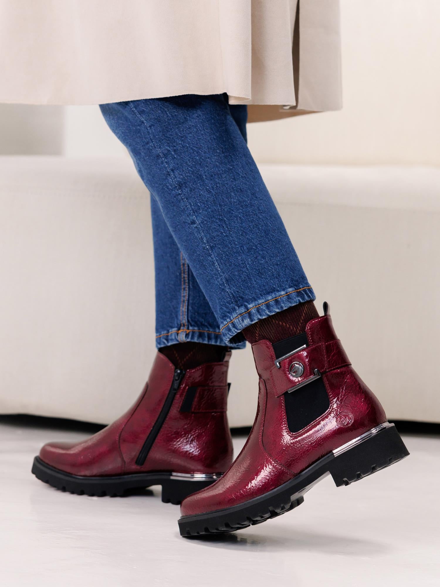 En person iført blå jeans, beige kåpe og skinnende rød 04676 Remonte Merano Ankle Boot med glidelås på siden, dekorativ spenne og uttagbar innersåle står på et lyst gulv i nærheten av en kremfarget sofa.