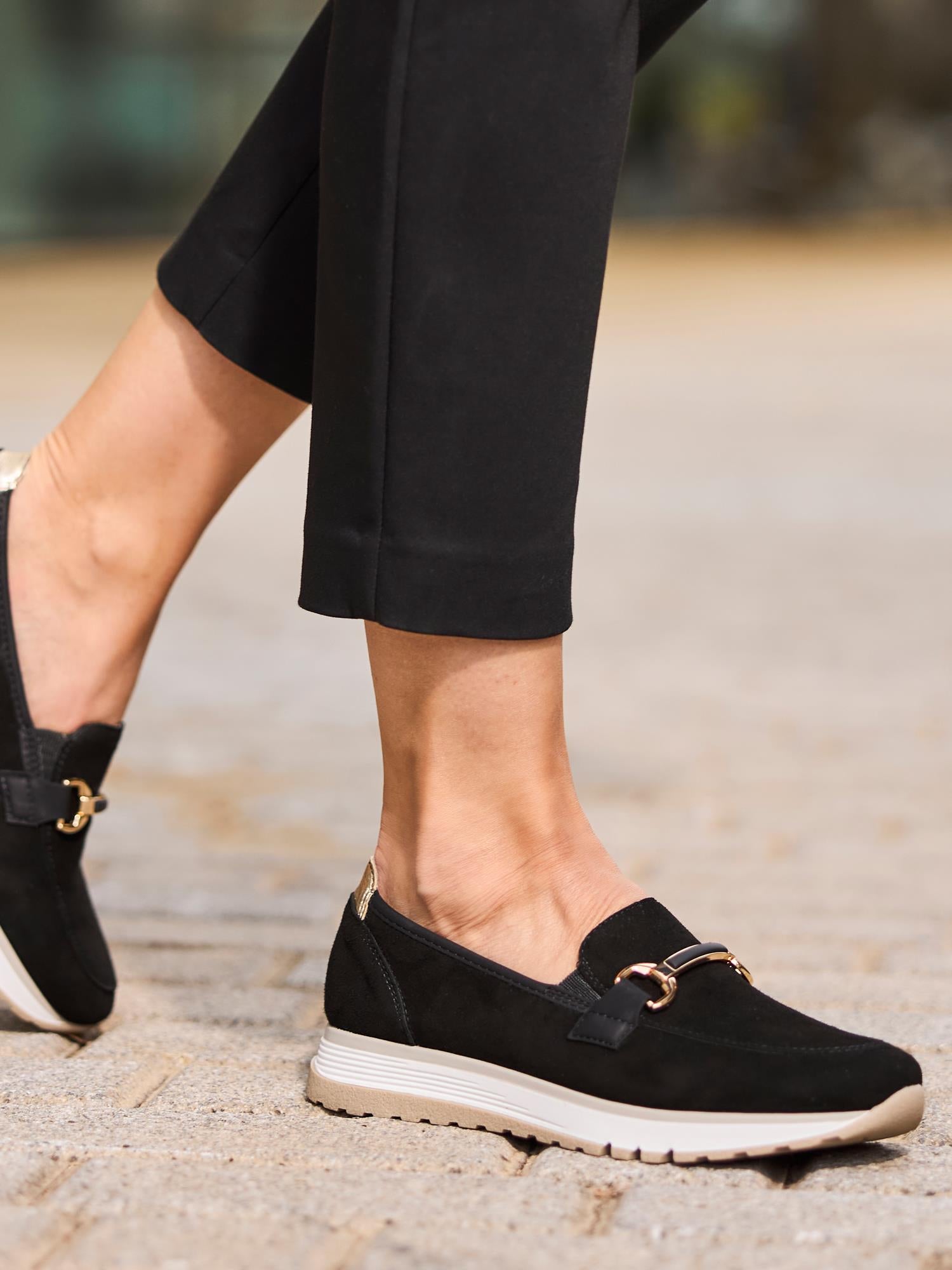 En person iført 02959 Rieker Soft Elegance Leather Black loafers fra Rieker Comfort & Style, med gulldetaljer og hvite såler, går på fortauet kledd i svarte ankellange bukser - perfekt for komfortskoentusiaster.