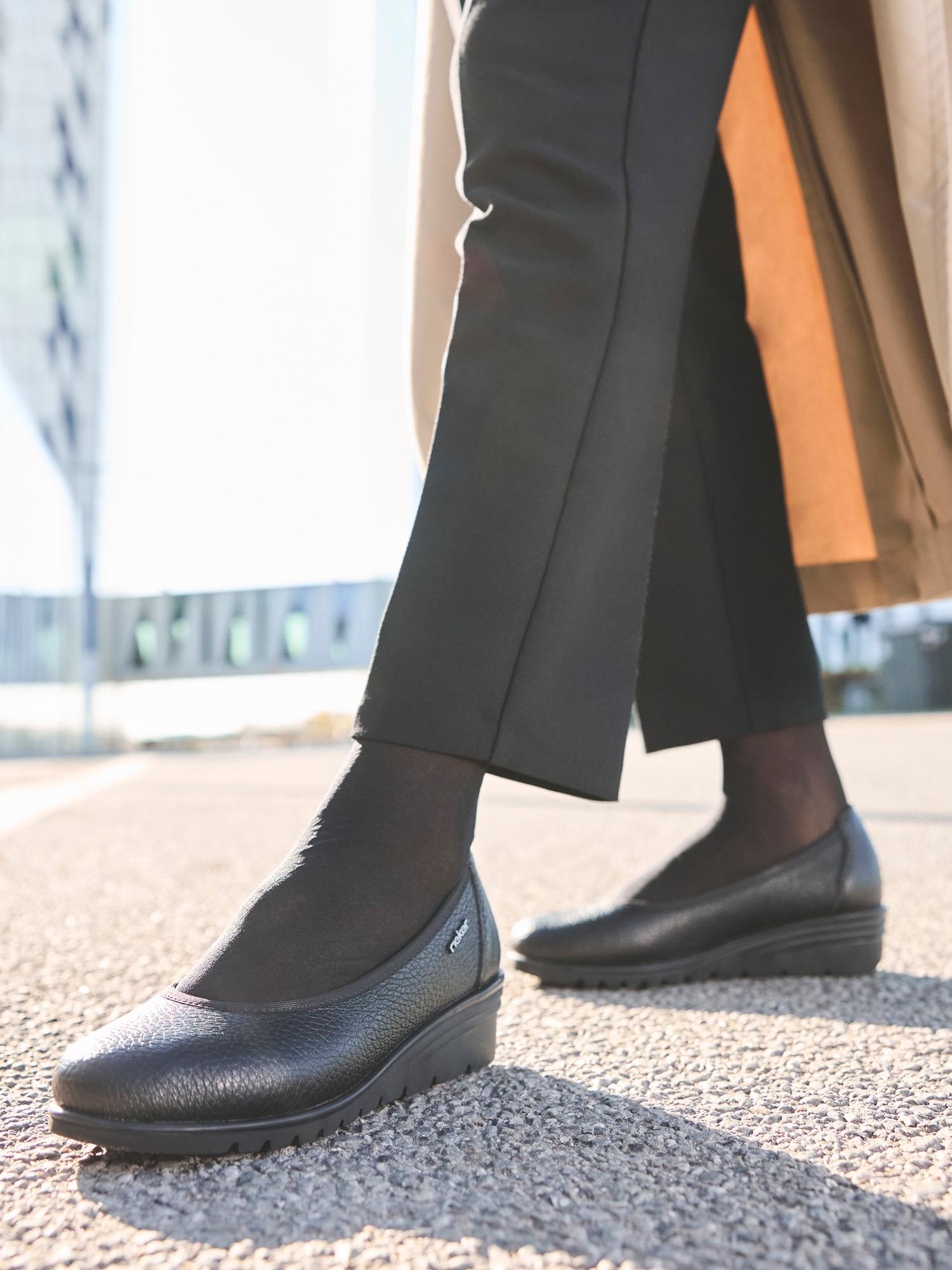 En person går utendørs på en solrik dag i svarte dressbukser, sorte, gjennomsiktige strømper og 04893 Rieker Liva Leather Ballerina fra Rieker Comfort & Style. Den nedre delen av en beige frakk vises mot en uskarp bybakgrunn.