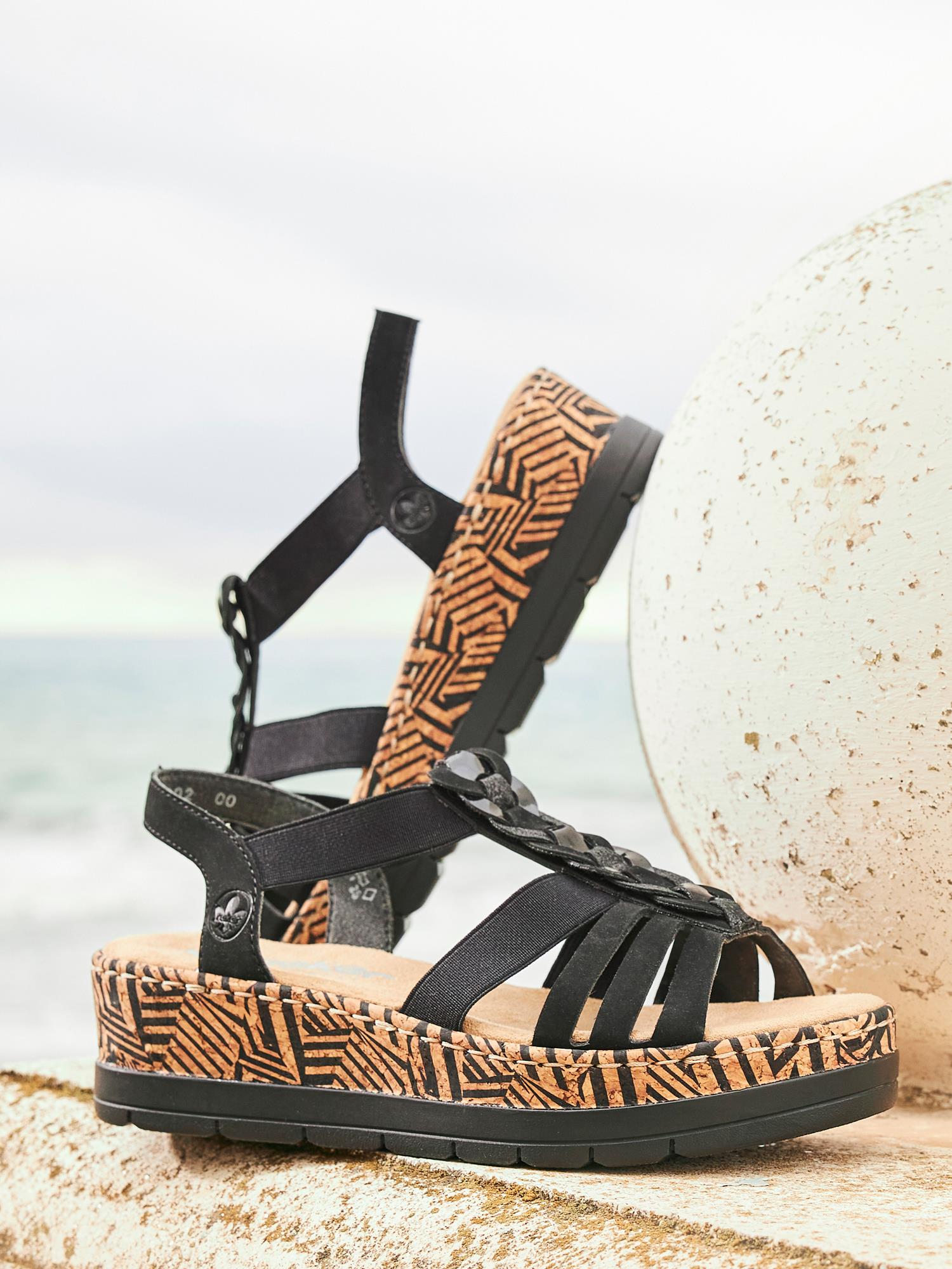 Et par 03370 Rieker Elviva Sandal fra Rieker Comfort & Style - svarte kilehælsandaler med stropper, såler med korkmønster og geometriske mønstre - vist utendørs i nærheten av en hvit kule og havet, elegante kilesandaler for maksimal komfort.