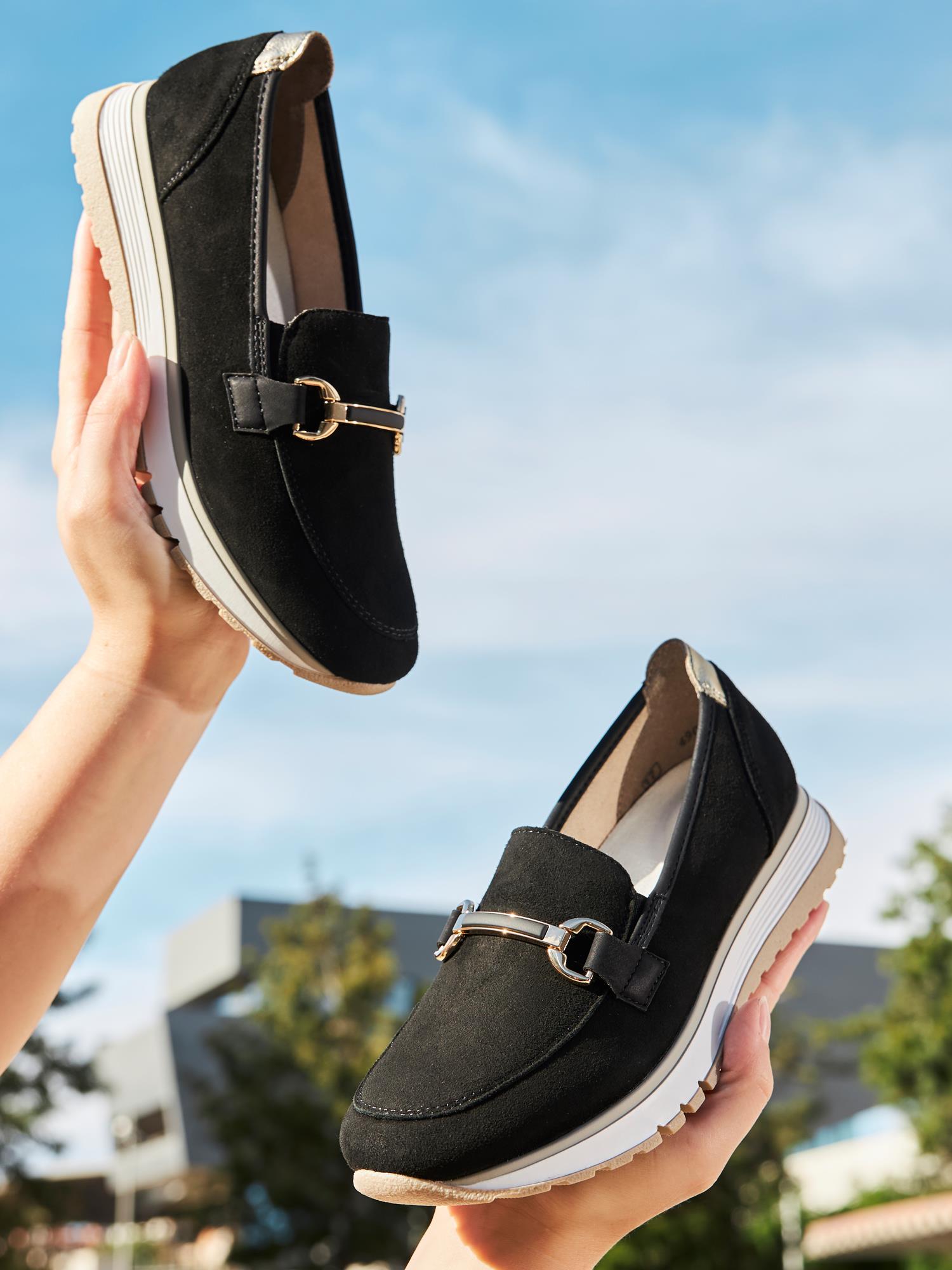 To hender holder opp et par 02959 Rieker Soft Elegance loafers i svart skinn med hvit såle og metalldetaljer mot en blå himmel - Rieker Comfort & Style sikrer komfort dame sko for stilig bruk hele dagen.