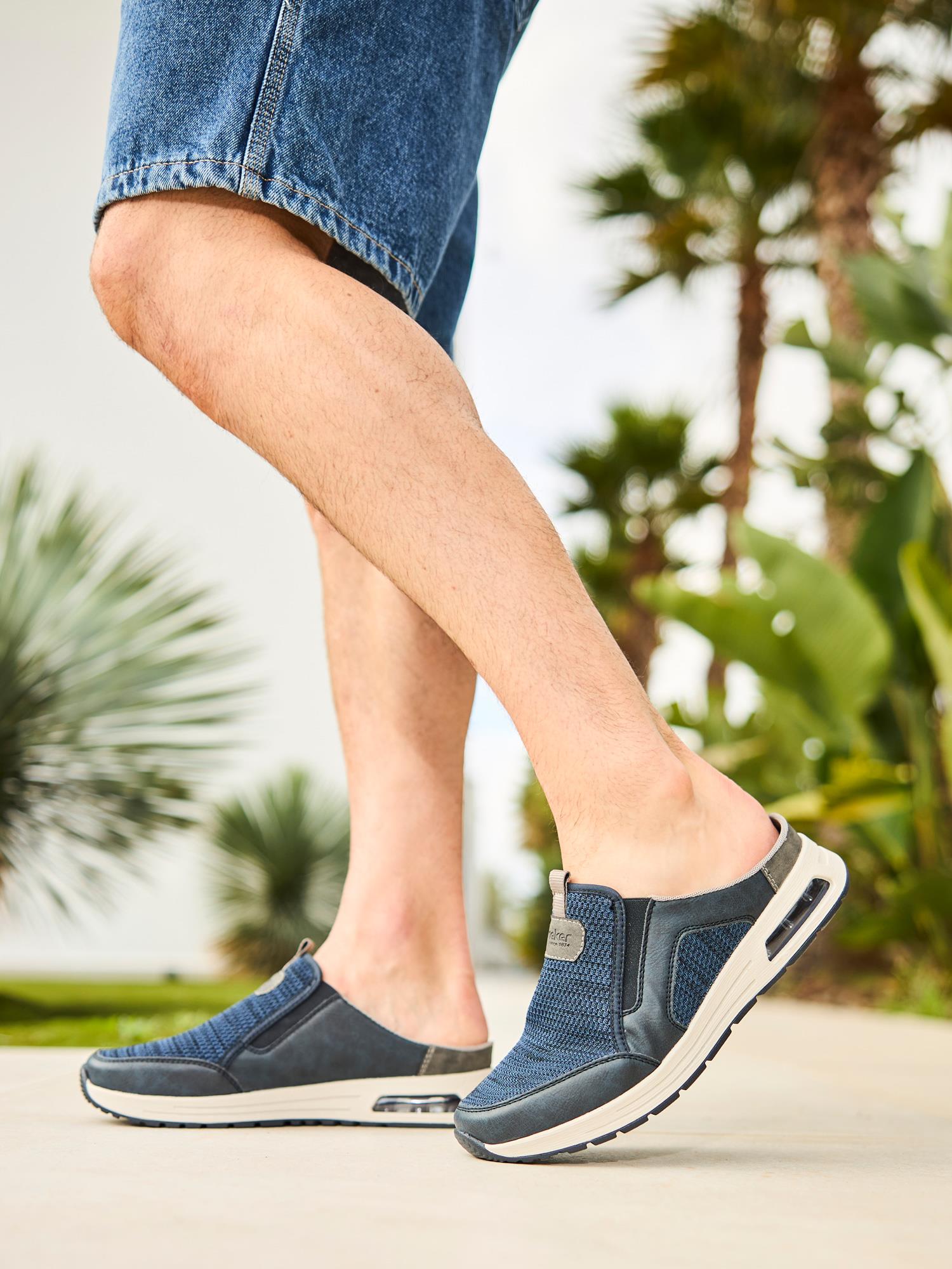 En person i blå 03051 Rieker Nolan Slip-on joggesko fra Rieker Comfort & Style og denimshorts går komfortabelt utendørs, omgitt av tropiske planter og palmer.