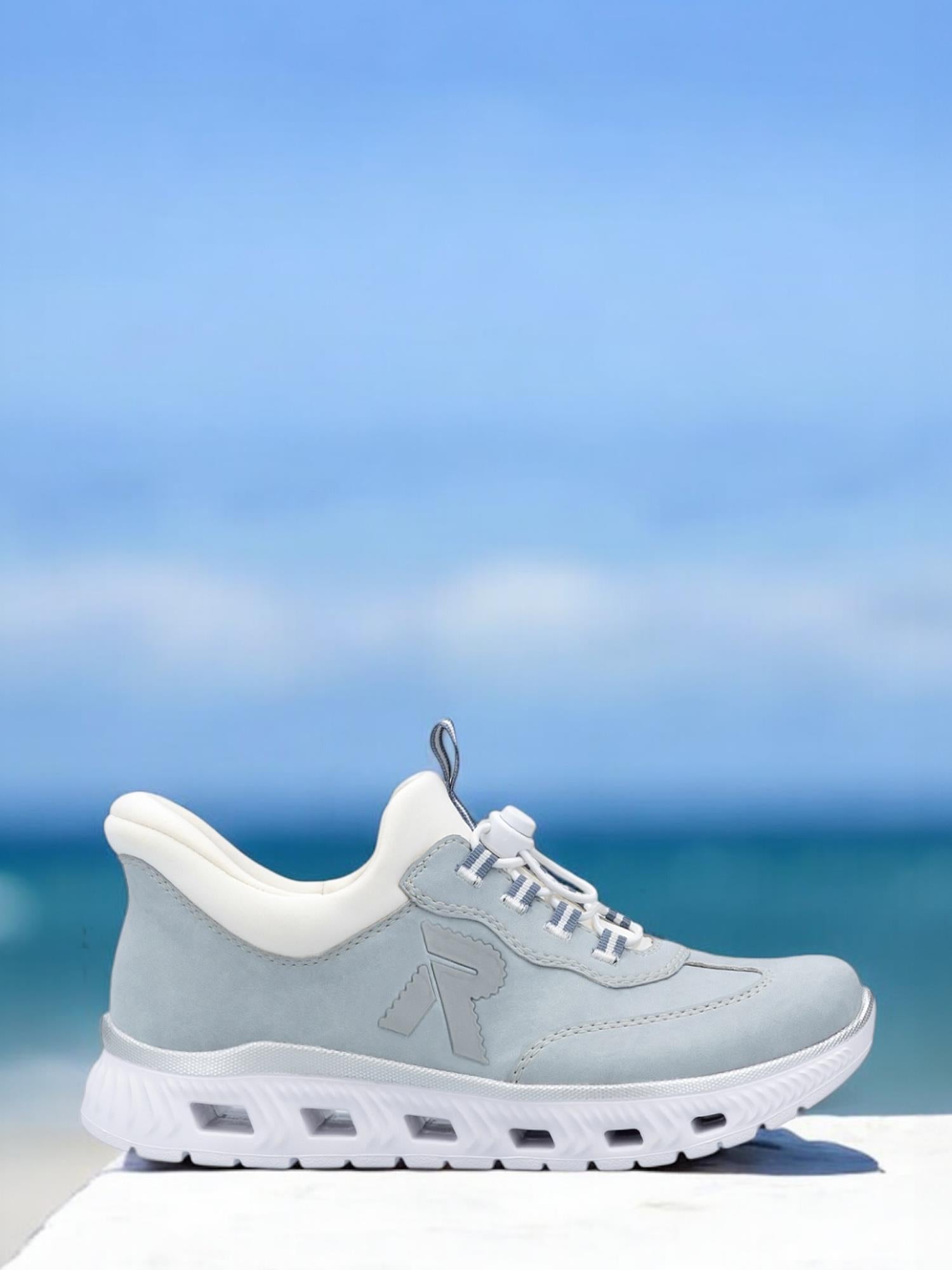 03278 Rieker Ready2GO Slip-On Sneakers fra Rieker Sport er avbildet utendørs på en hvit flate, med en uskarp blå himmel og havet bak. Disse lyseblå og hvite joggeskoene har en tykk hvit såle og snørelukking for komfort.