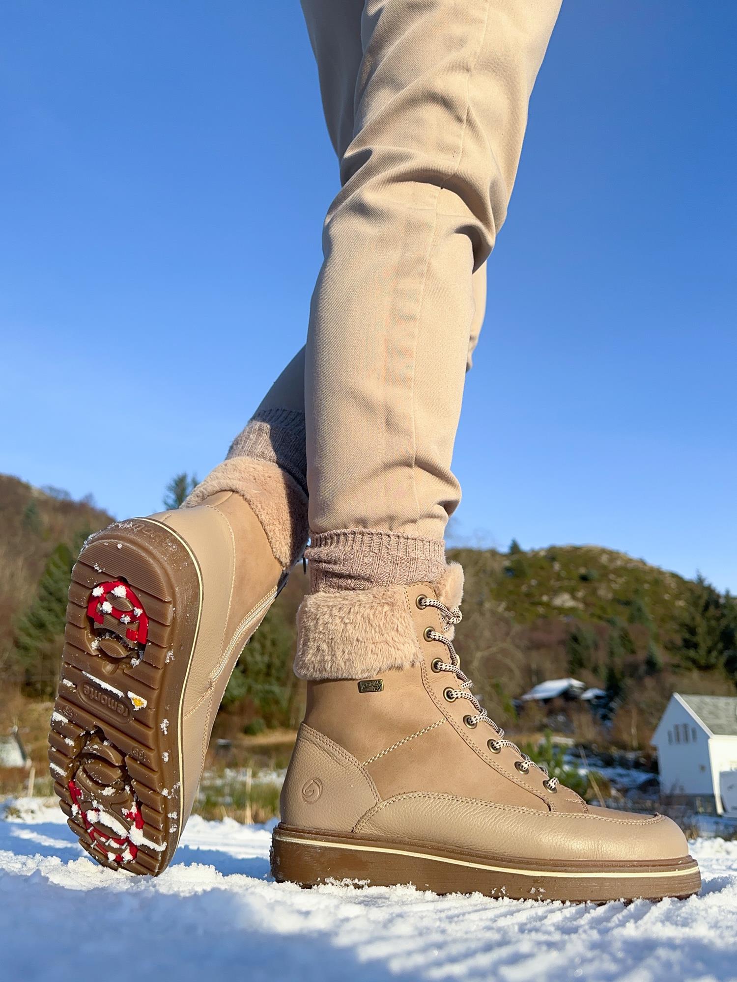 En person i beige bukser står på snøen og viser frem sålen med rødt og hvitt grep på Remonte 04803 Premium leather tex nude ullfôret med brodder boots. I bakgrunnen skimtes trær og hus under en klar himmel.