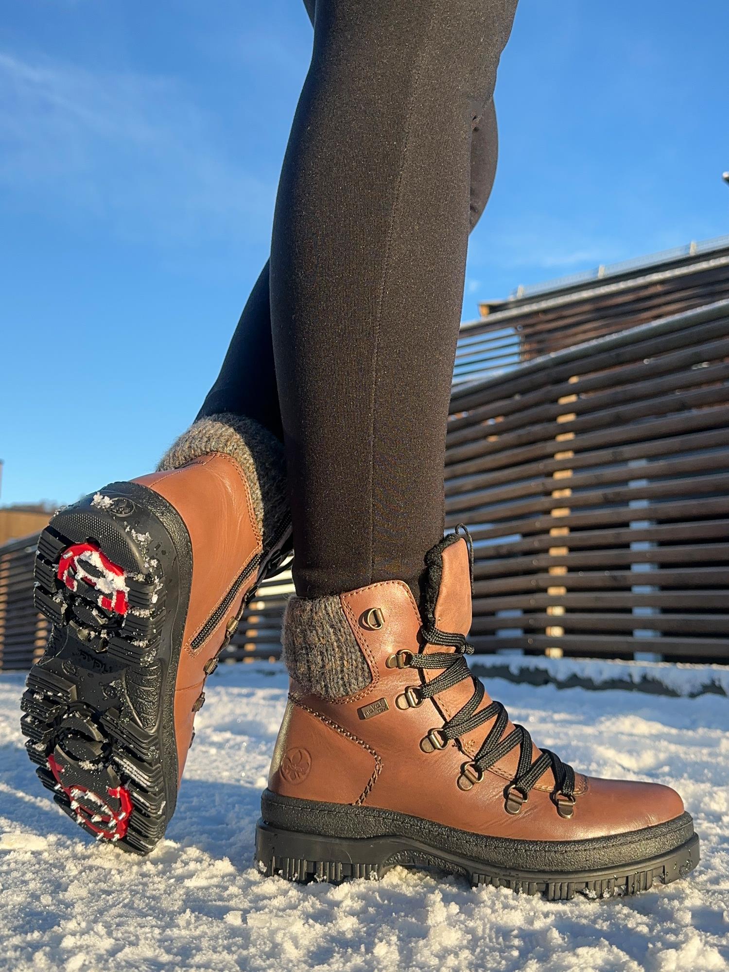 En person i svarte leggings og 05038 Rieker Leather tex varmfôret med brodder fra Rieker Comfort & Style står på snø foran en moderne trebygning. De burgunderrøde støvlene har tykke såler, brodder for grep og et vanntett RiekerTEX-membran.