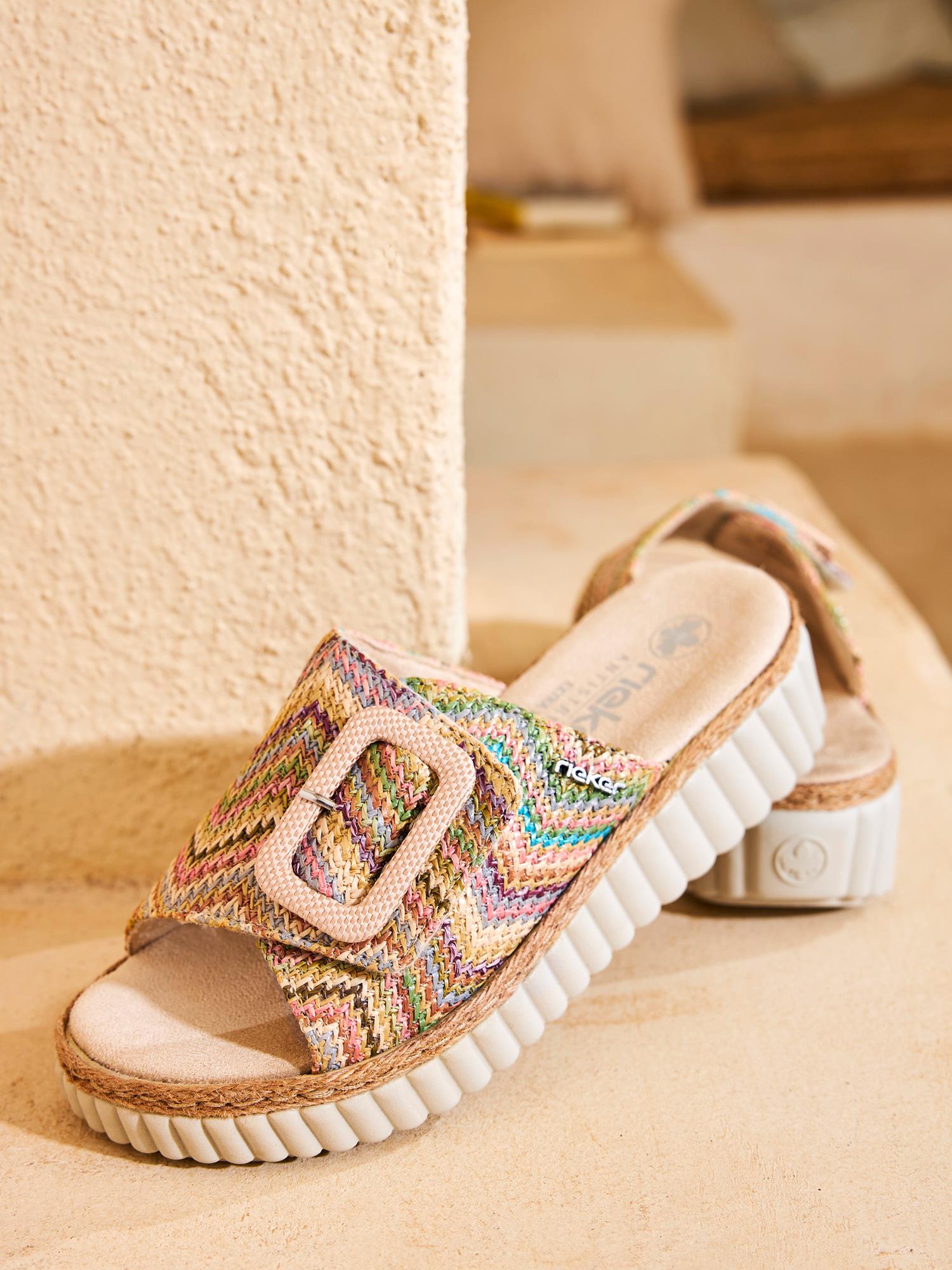 03049 Rieker Solenya Slip-in Sandal fra Rieker Comfort & Style har vevd, flerfarget overdel, tykke, hvite, riflede såler og en stor spenne, som vises mot en strukturert beige vegg i mykt innelys for ultralett komfort.