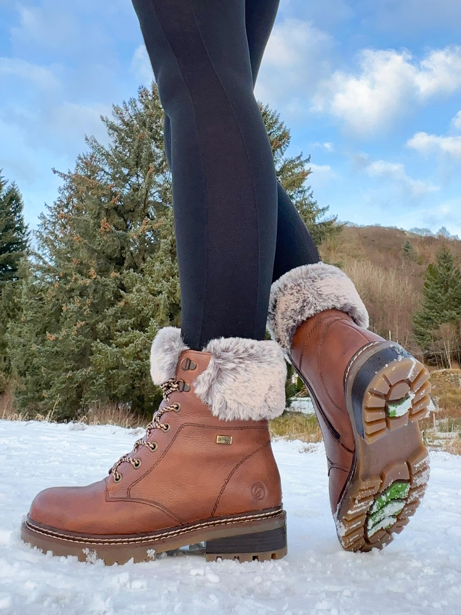 En person i svarte leggings står på snødekt mark, iført Remonte 04801 vintersko skinn ullfôret fibergripstøvler med brunt pelsfôr, synlige slitebaner og metalløyer; i bakgrunnen ser man eviggrønne trær og blå himmel.