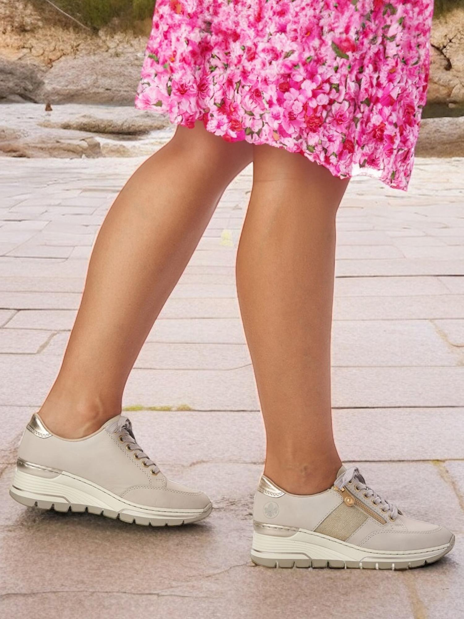 En person i et rosa blomstrete skjørt og 06829 Rieker Golden Beige Sneakers fra Rieker Comfort & Style går på et steinbelegg, med bare bena og føttene synlige.