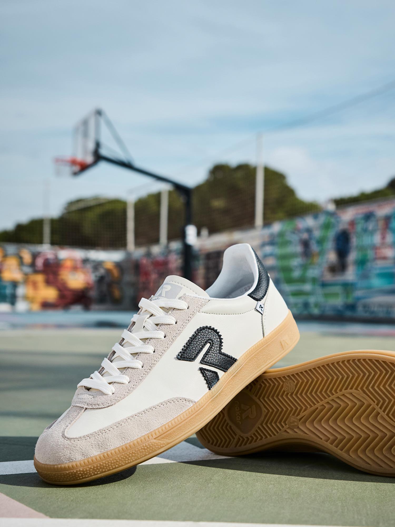 03337 Rieker San Diego Leather Sneaker White fra Rieker Sport, med svart "R"-logo og Swing2Go-teknologi for komfort, er avbildet på en utendørs basketballbane med en basketballkurv og levende veggmalerier i bakgrunnen.