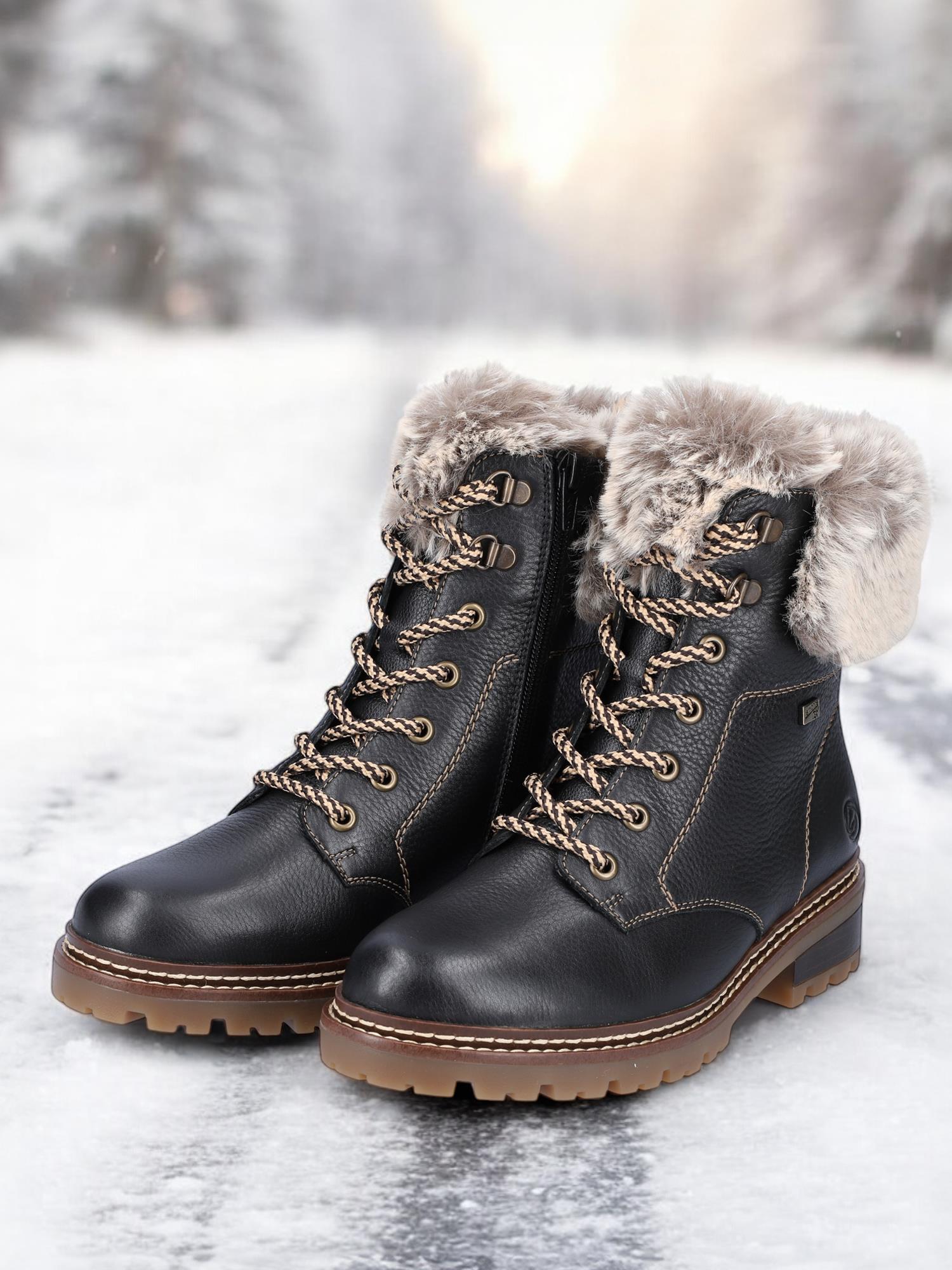 Et par svarte Remonte 04794 vintersko skinn ullfôret med Fiber-Grip solbrune såler er plassert på en snødekt sti, med uskarpe trær i bakgrunnen.