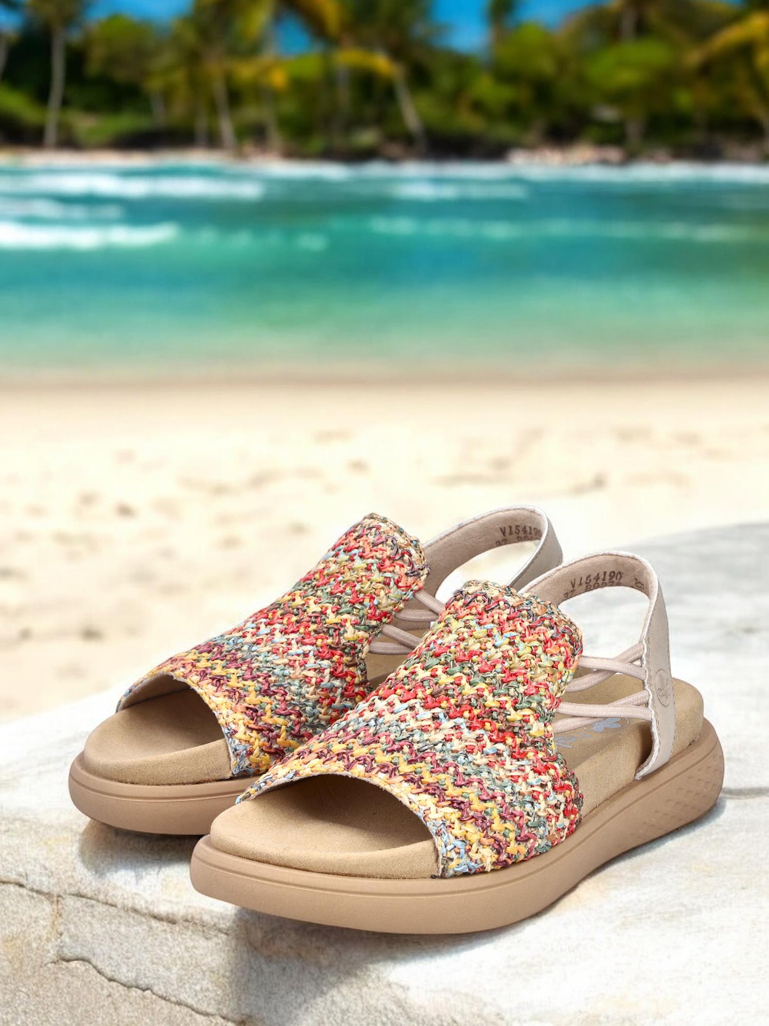Et par 03341 Rieker Viala Sandal fra Rieker Comfort & Style med fargerik komfort hviler på en hvit stein, mot en uskarp tropisk strand, turkist hav og palmer i bakgrunnen.