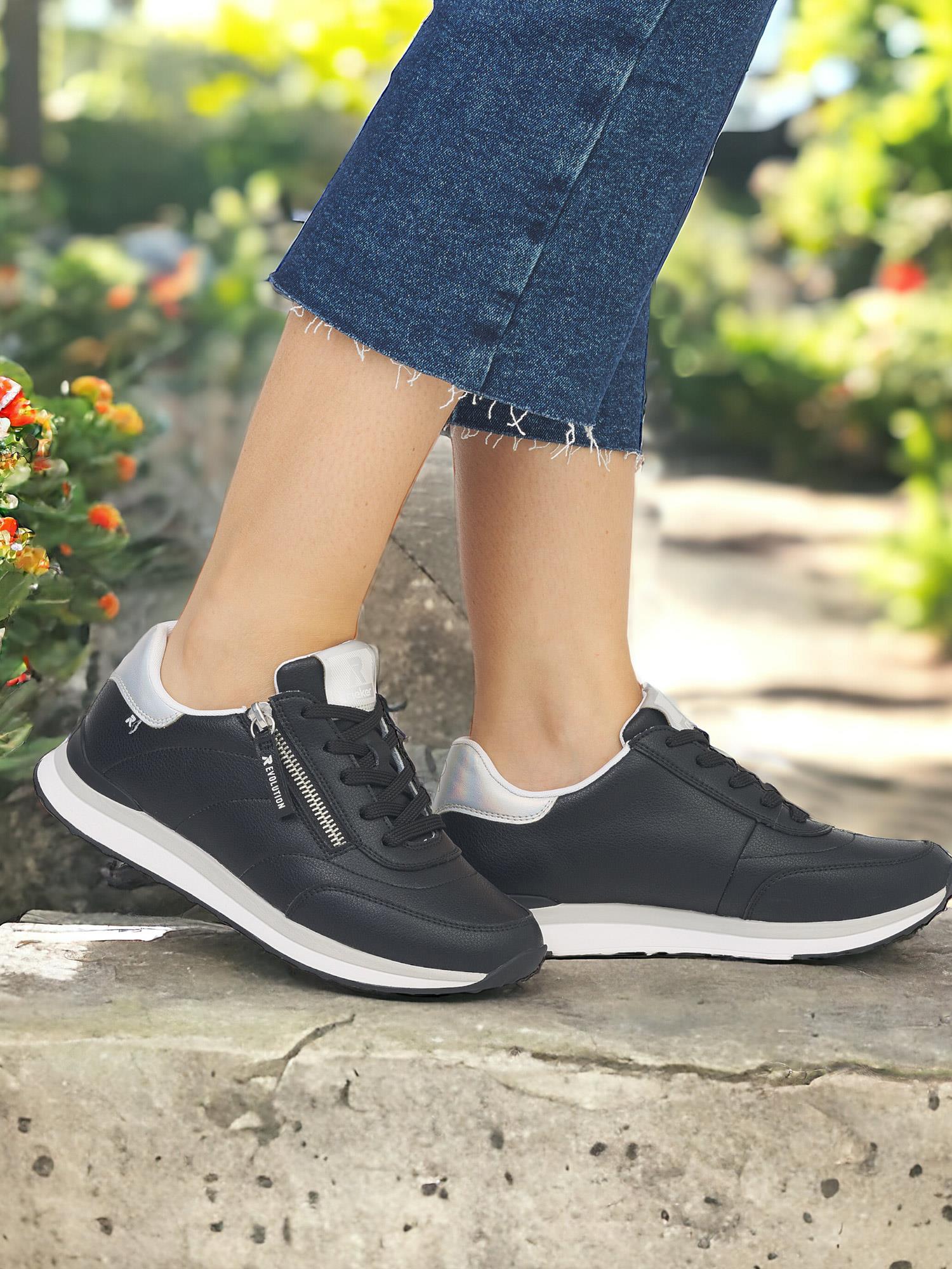 En person i korte blå jeans står på en steinsti blant grøntområder og blomster, iført svarte Rieker Sport 07537 Rieker-Evolution Memory Sneaker skinnsko med hvite såler og sølvdetaljer.