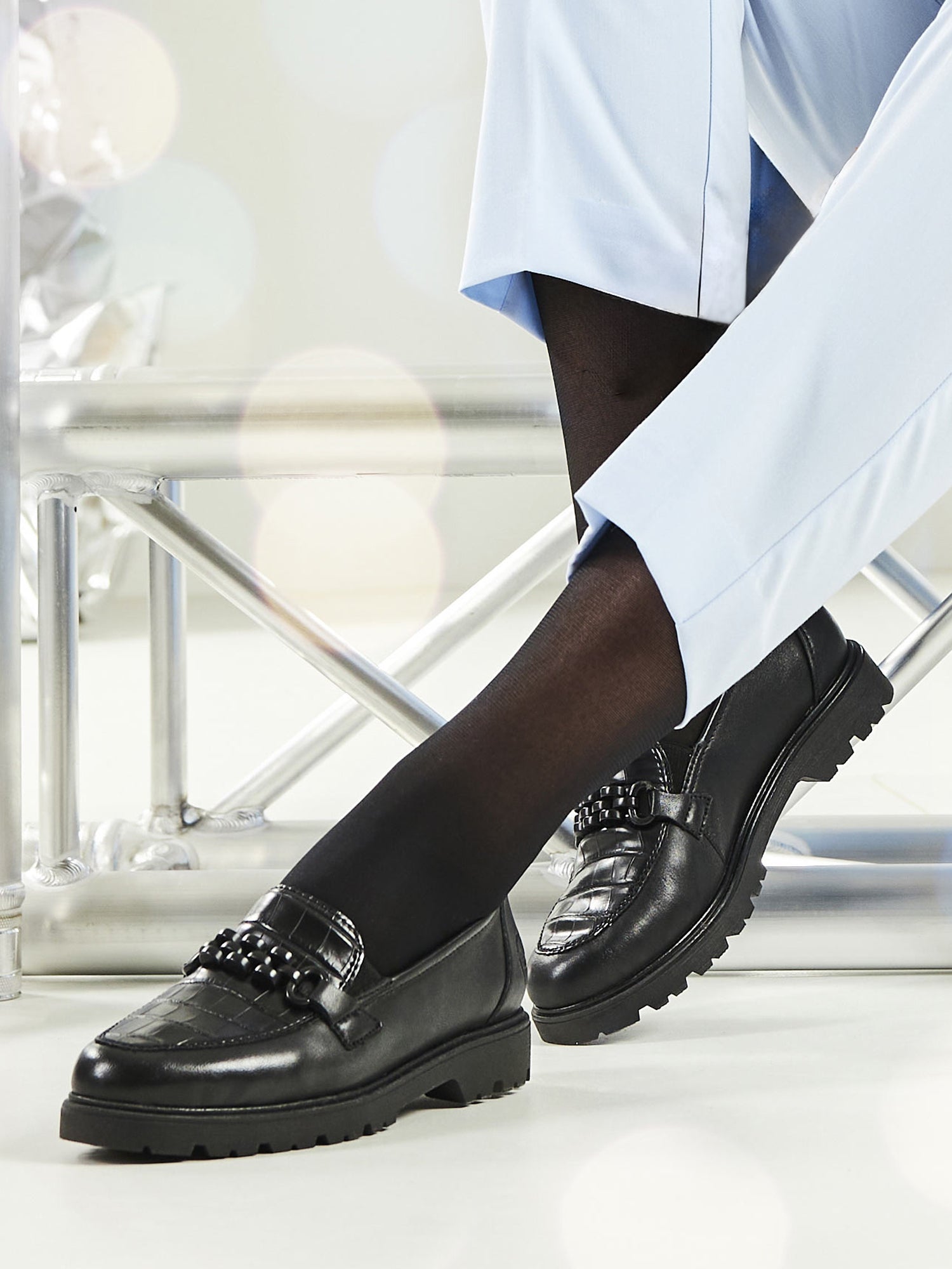 En person iført lyseblå bukser, svarte strømpebukser og Rieker Comfort & Style 04737 Palma Leather Loafers med dekorative nitter sitter på en moderne metallstol. Fokuset er på bena og de behagelige skoene mot en mykt uskarp bakgrunn.