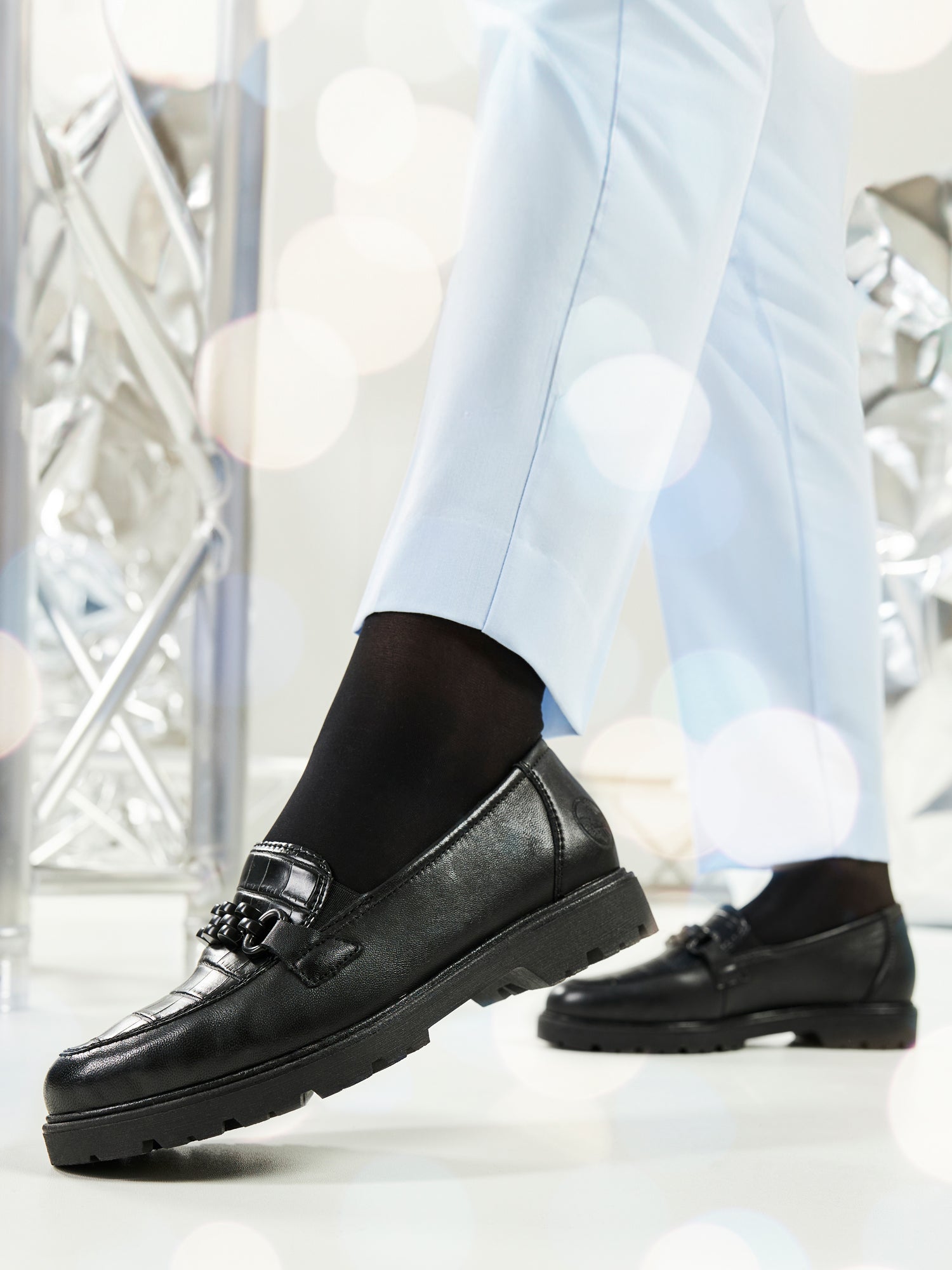 En person innendørs har på seg lyseblå bukser, svarte sokker og 04737 Rieker Palma Leather Loafer fra Rieker Comfort & Style med dekorative dusker; metalliske strukturer og myke lysbokeh vises i bakgrunnen.