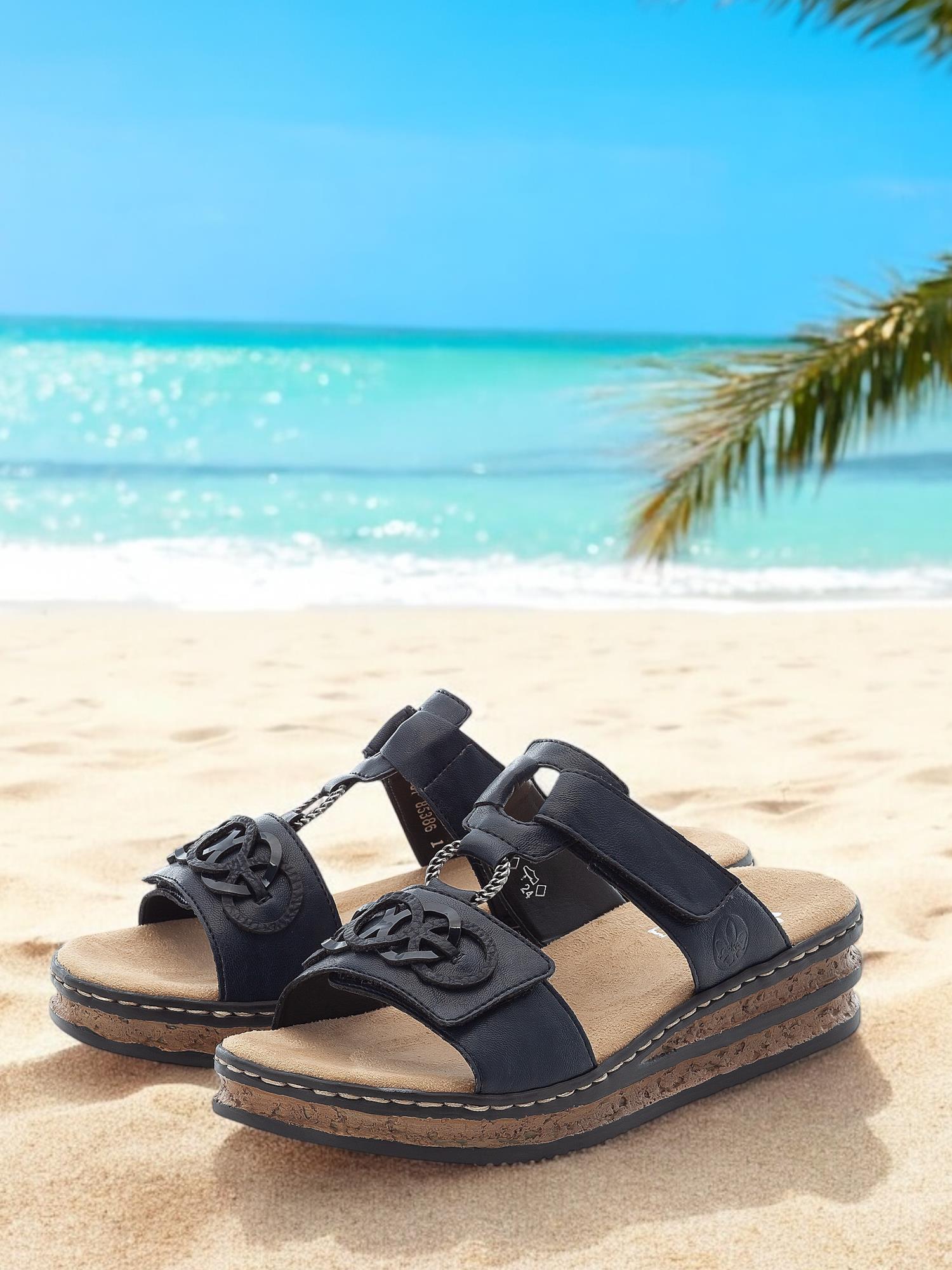 02996 Rieker Pacific Dream-sandalen fra Rieker Comfort & Style med komfort og korkdetaljer hviler på en sandstrand nær palmeblader, innrammet av turkis hav og klarblå himmel.