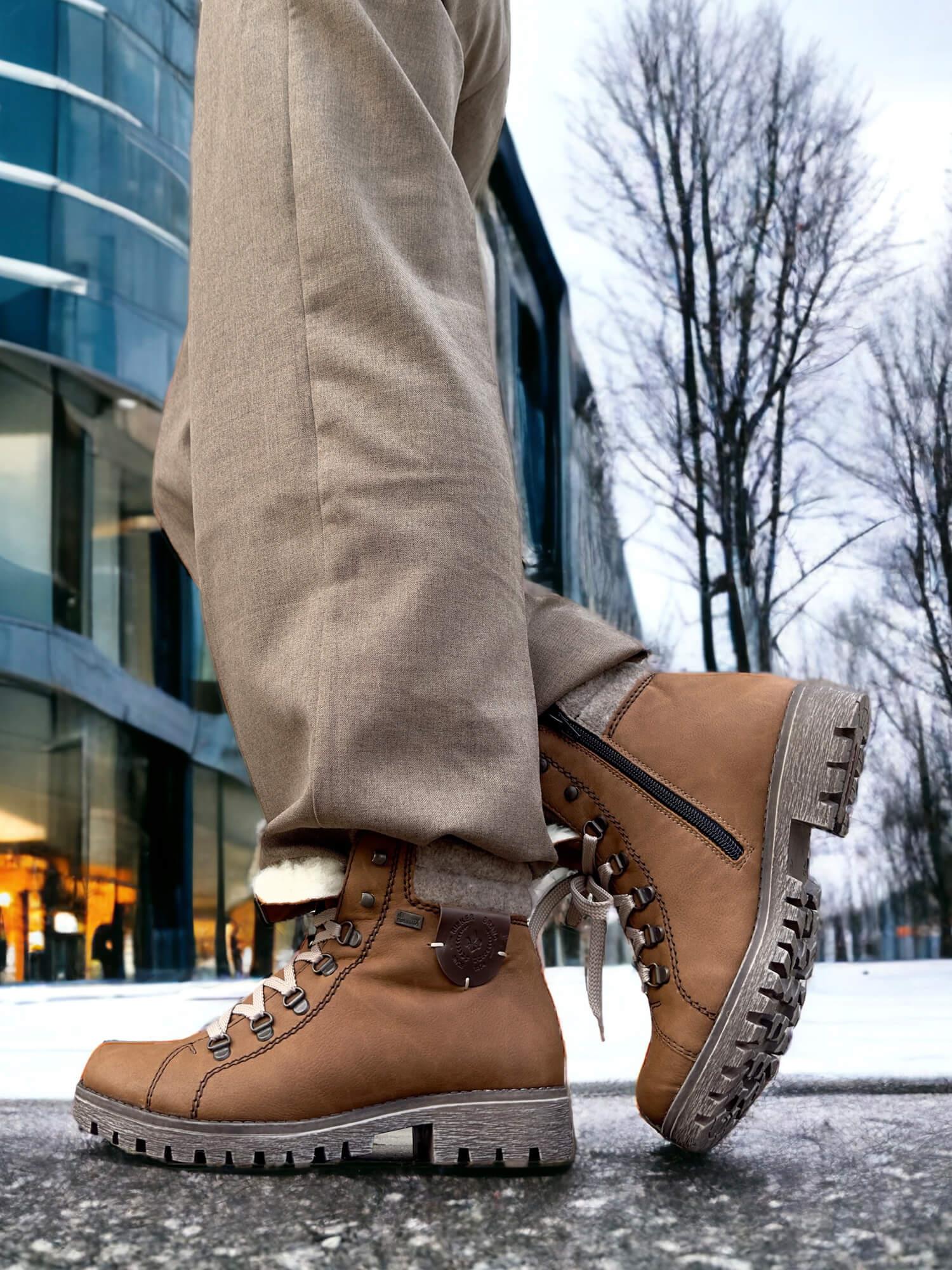 En person iført Rieker Comfort & Style 03043 Varmfòret Vintersko ULL brune og beige bukser står utendørs på et snødekt fortau, med moderne glassbygninger og bladløse trær i bakgrunnen.