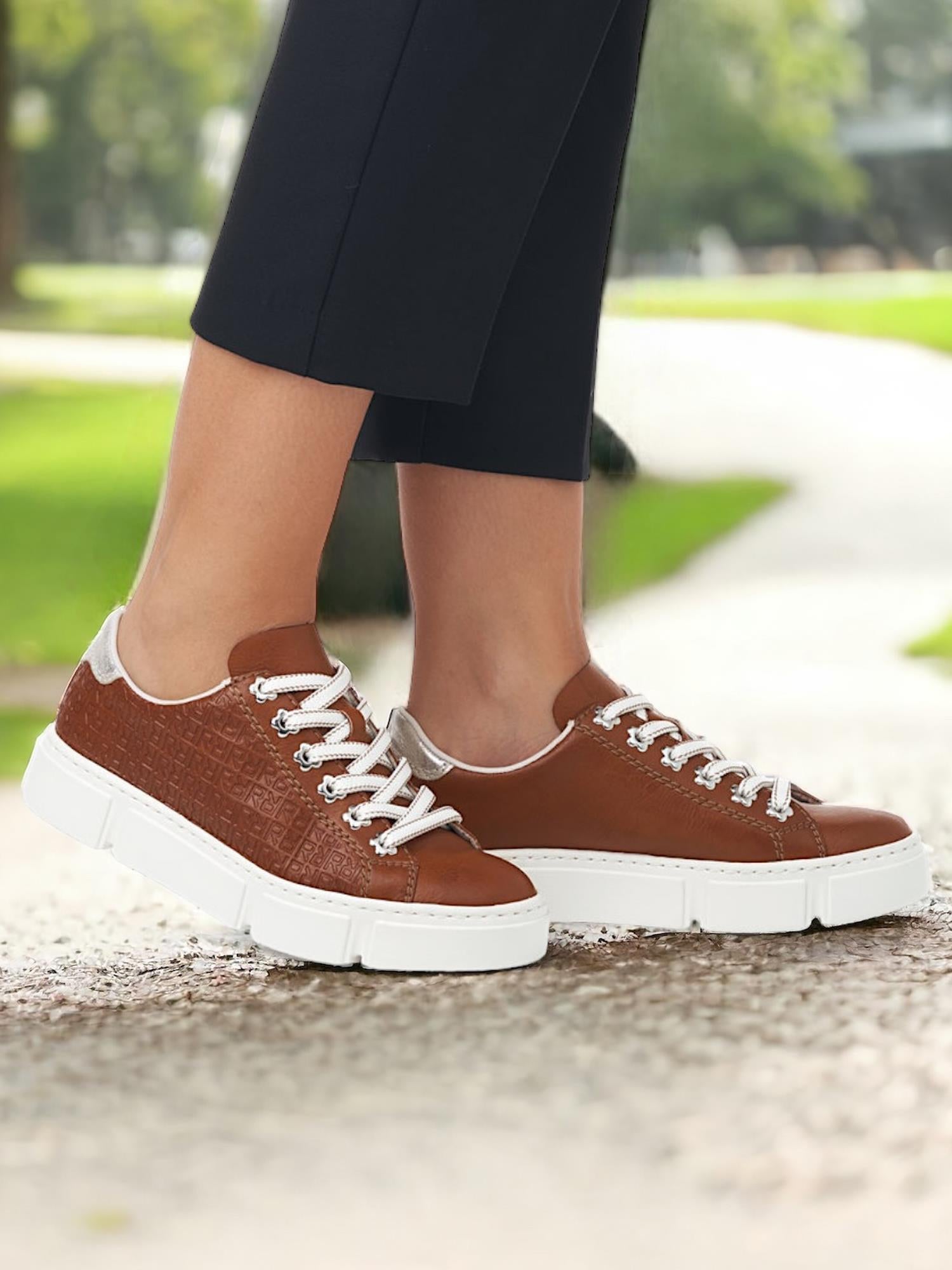 Noen står utendørs på en sti iført Rieker Comfort & Style 07575 Tawny Leather Sneakers med hvite såler og lisser, med grønt gress og trær mykt uskarpt i bakgrunnen.