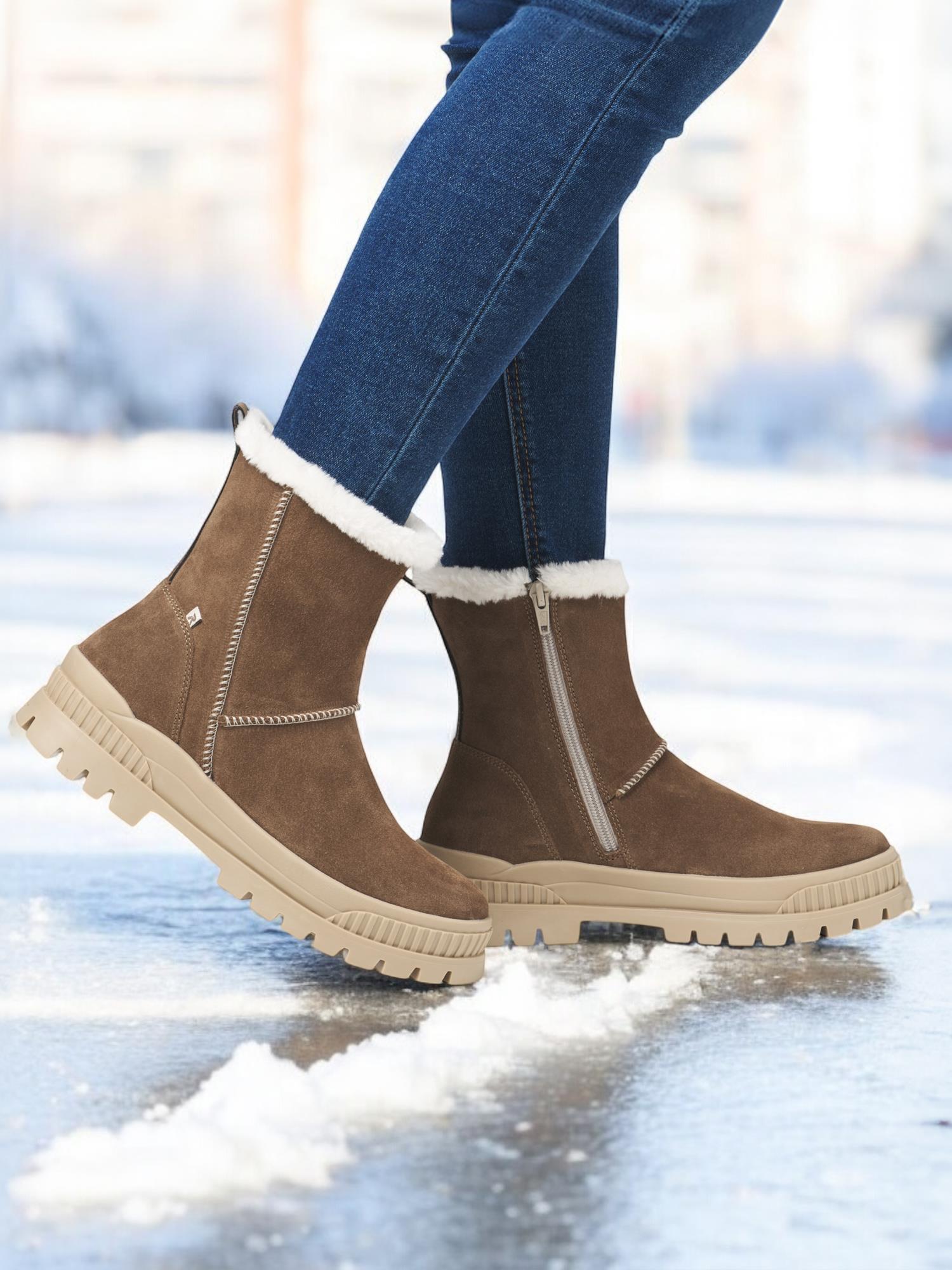En person iført Rieker Sport 05069 Rieker-Evolution Nubuck Ullfôret Fiber-Grip vintersko med beige såler og hvitt pelsfôr står på snø, de brune støvlene med glidelåser på siden er satt sammen med blå skinny jeans.