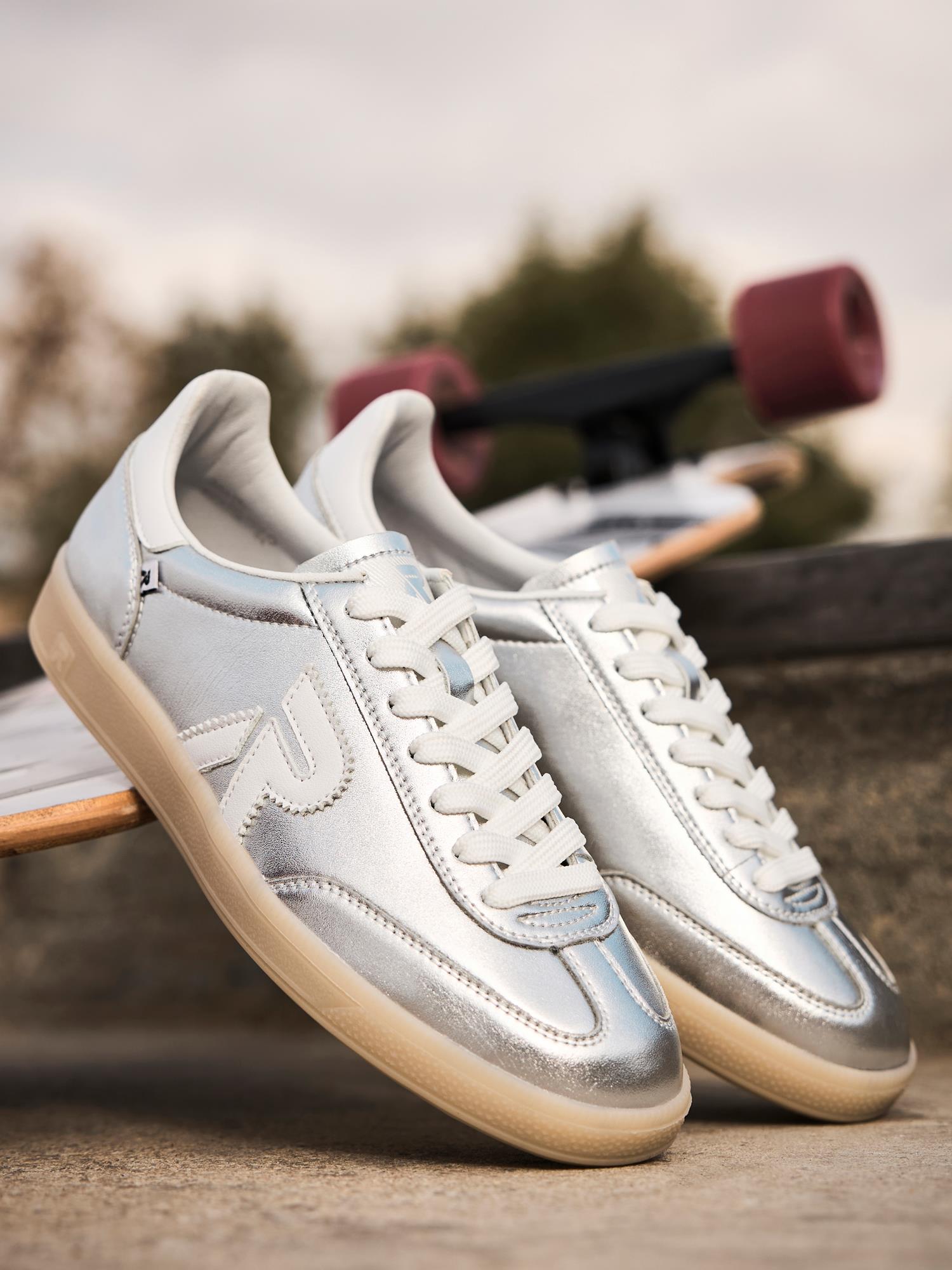 Et par 03524 Rieker Rieker San Diego Leather Sneaker Silver fra Rieker Sport med hvite lisser står på et skateboard utendørs, mot en uskarp bakgrunn av trær og en overskyet himmel.