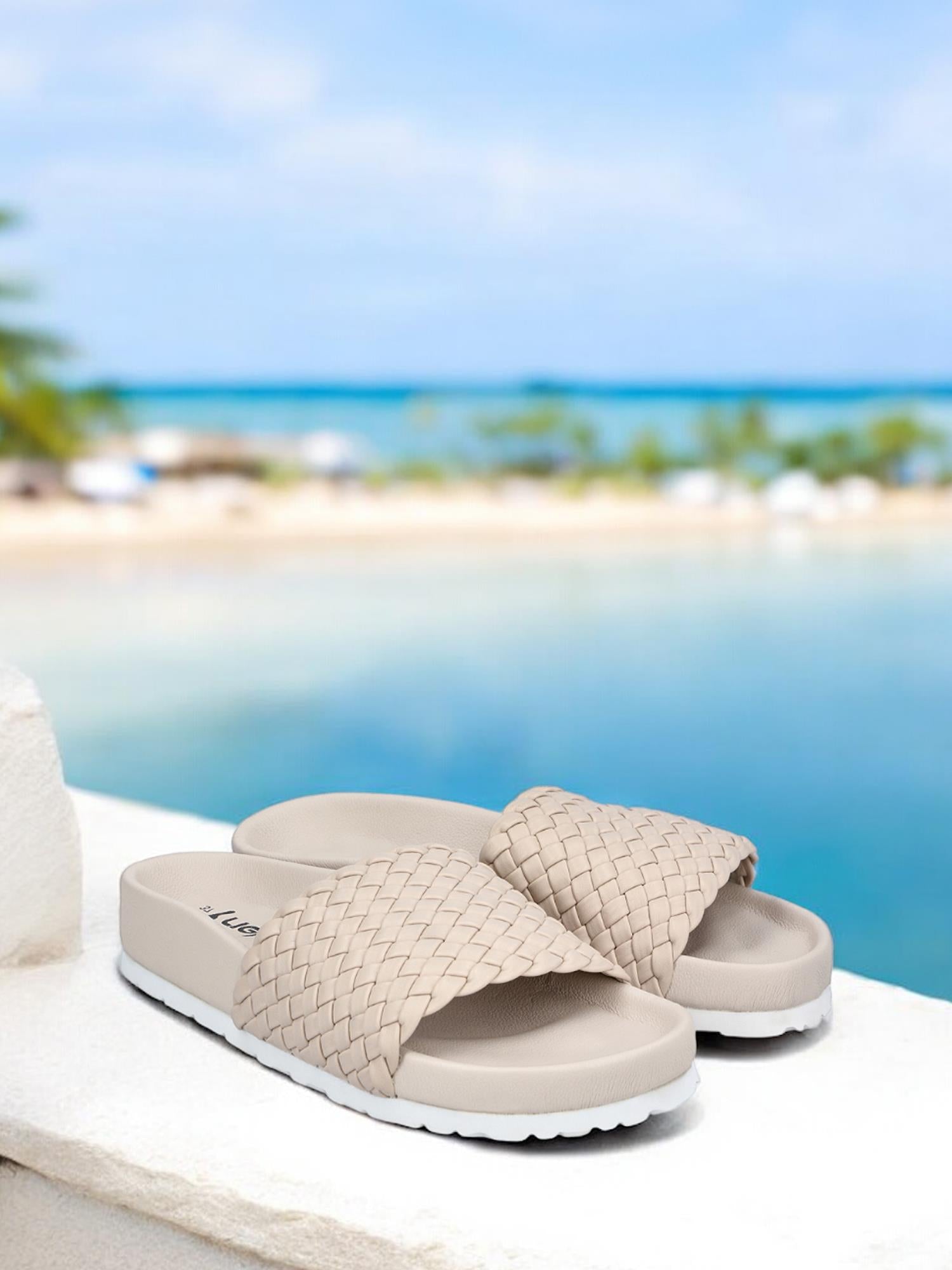 08093 Rieker Ginger Braid Sandal fra Rieker Comfort & Style, i beige med hvit såle, har komfort- og antistress-teknologi, avbildet på en hvit avsats mot en tropisk strand med blått vann og palmer.