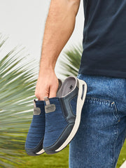 Iført blå jeans og mørk skjorte holder en person de blå 03051 Rieker Nolan Slip-on joggeskoene fra Rieker Comfort & Style med hvite såler; uskarpe grønne planter bak fremhever komfort og stil.