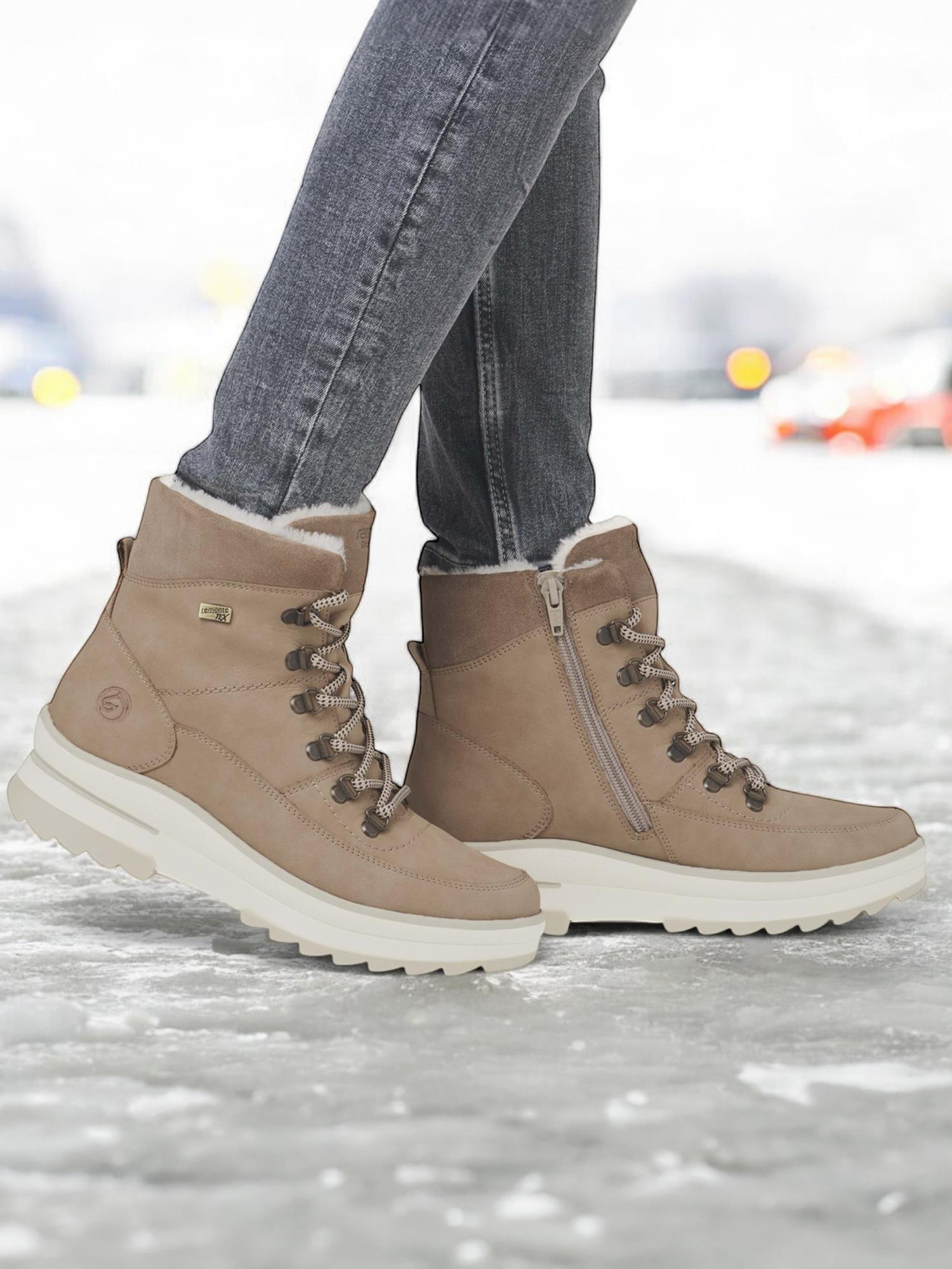 En person iført Remonte 05022 Fiber-Grip tex-membran skinn vinterstøvler med ullfôr og tykke, hvite såler står på snøen i grå dongeribukser. Bakgrunnen er uskarp med antydninger til kjøretøy og lys.