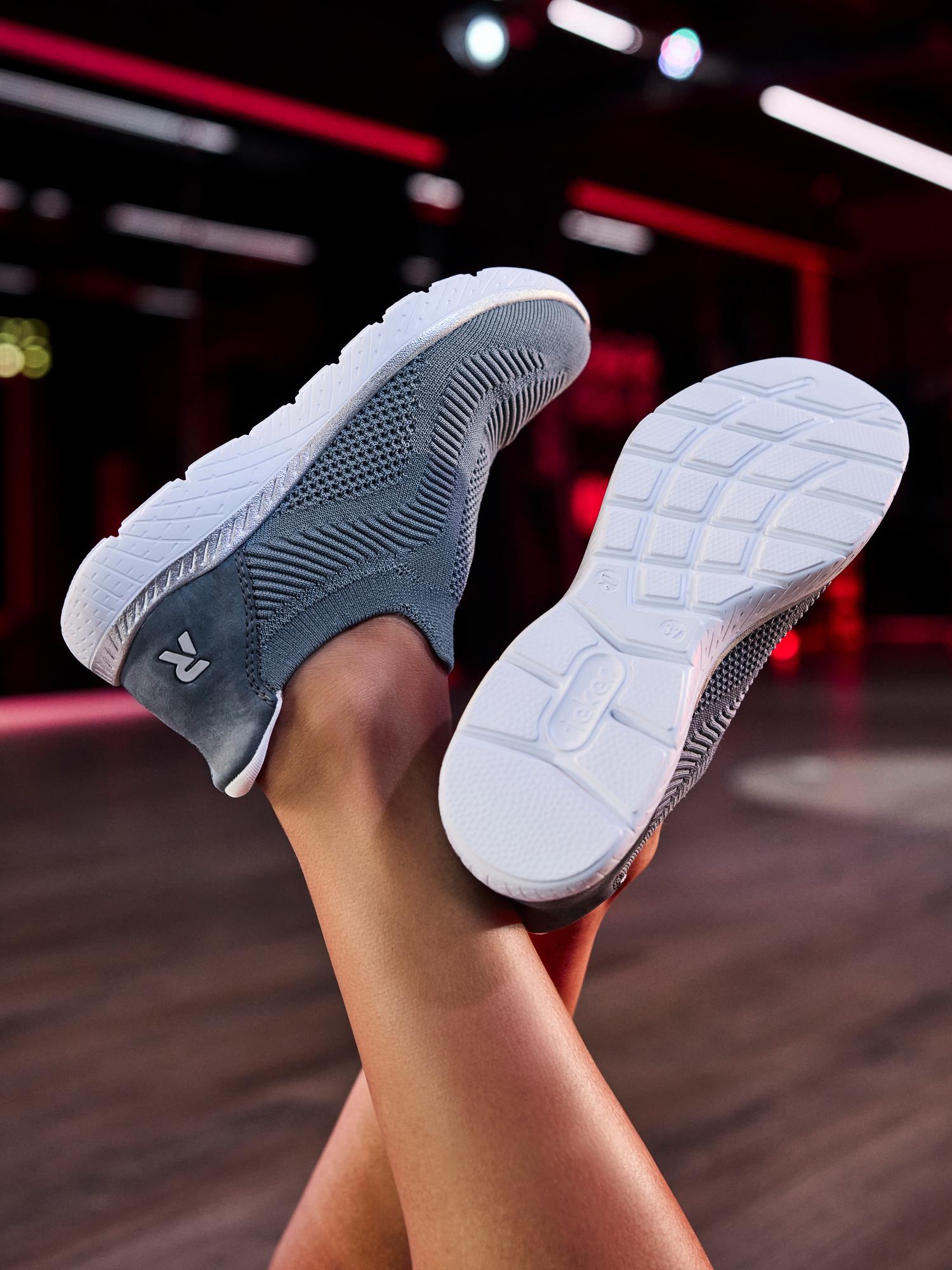 En persons ben er krysset, og avslører blå Rieker Sport 07136 Ready2GO slip-on joggesko med hvite såler og strikket tekstur, med logoen på hælen. Innendørsmiljøet har dempet, fargerik belysning for komfort hele dagen.