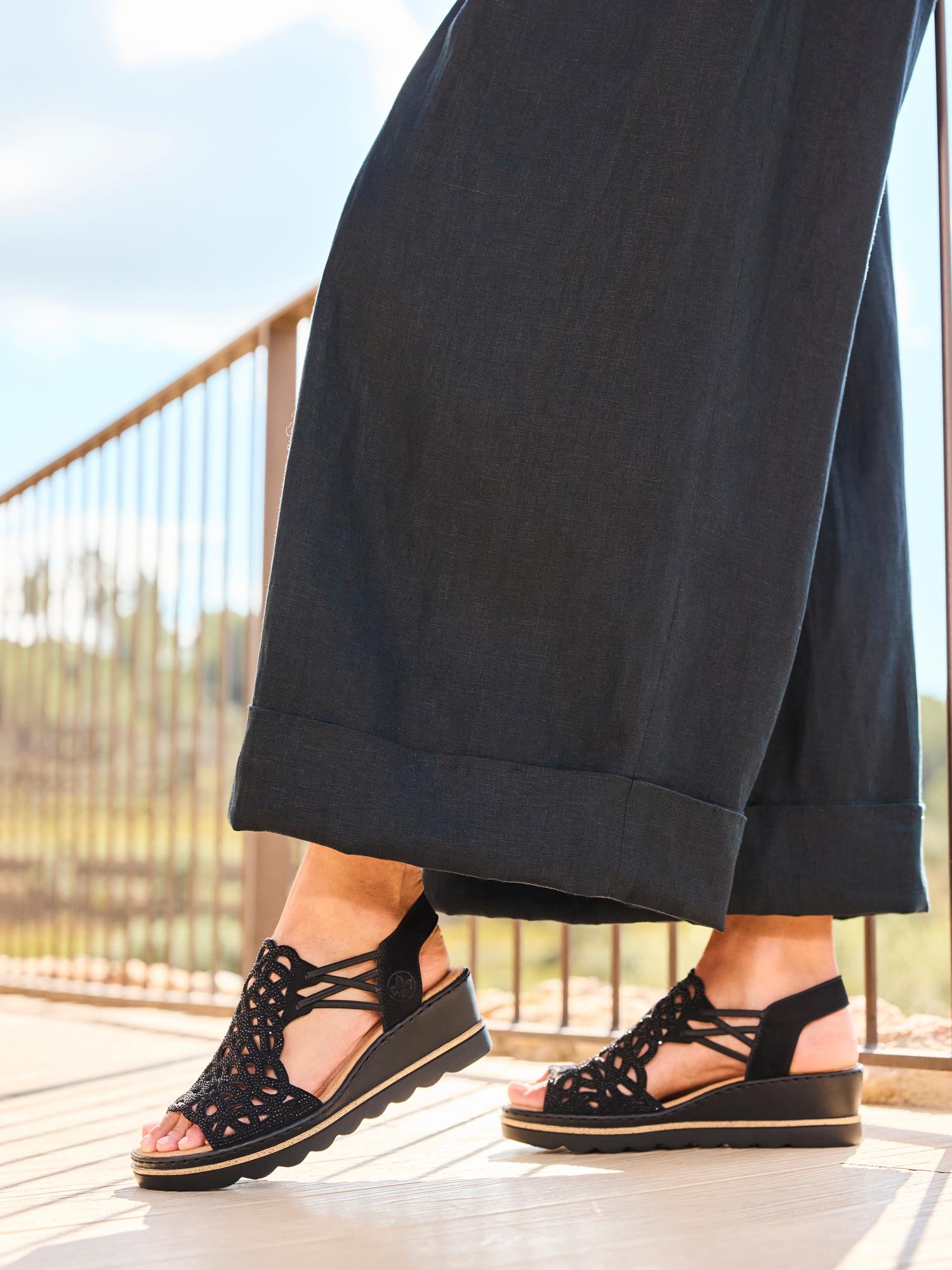 Iført sorte bukser med vide ben og 03003 Rieker Sorella Sandal fra Rieker Comfort & Style står en person utendørs på en solbelyst uteplass med et metallrekkverk og et uskarpt grønt landskap i bakgrunnen.