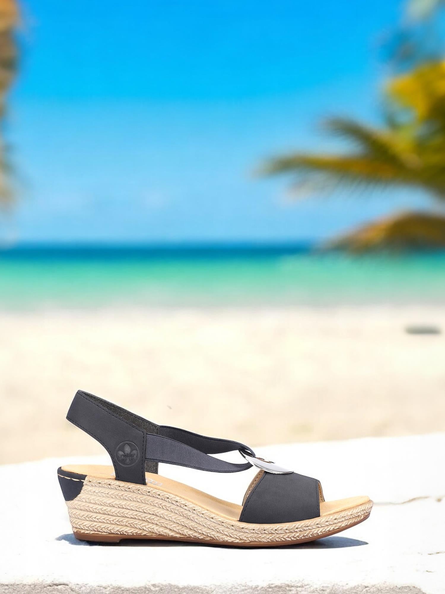 08079 Rieker Pazifik Elastic Sandal fra Rieker Comfort & Style, i svart med vevd såle, er avbildet på en hvit flate med en uskarp tropisk strand, turkis hav og palmer i bakgrunnen.