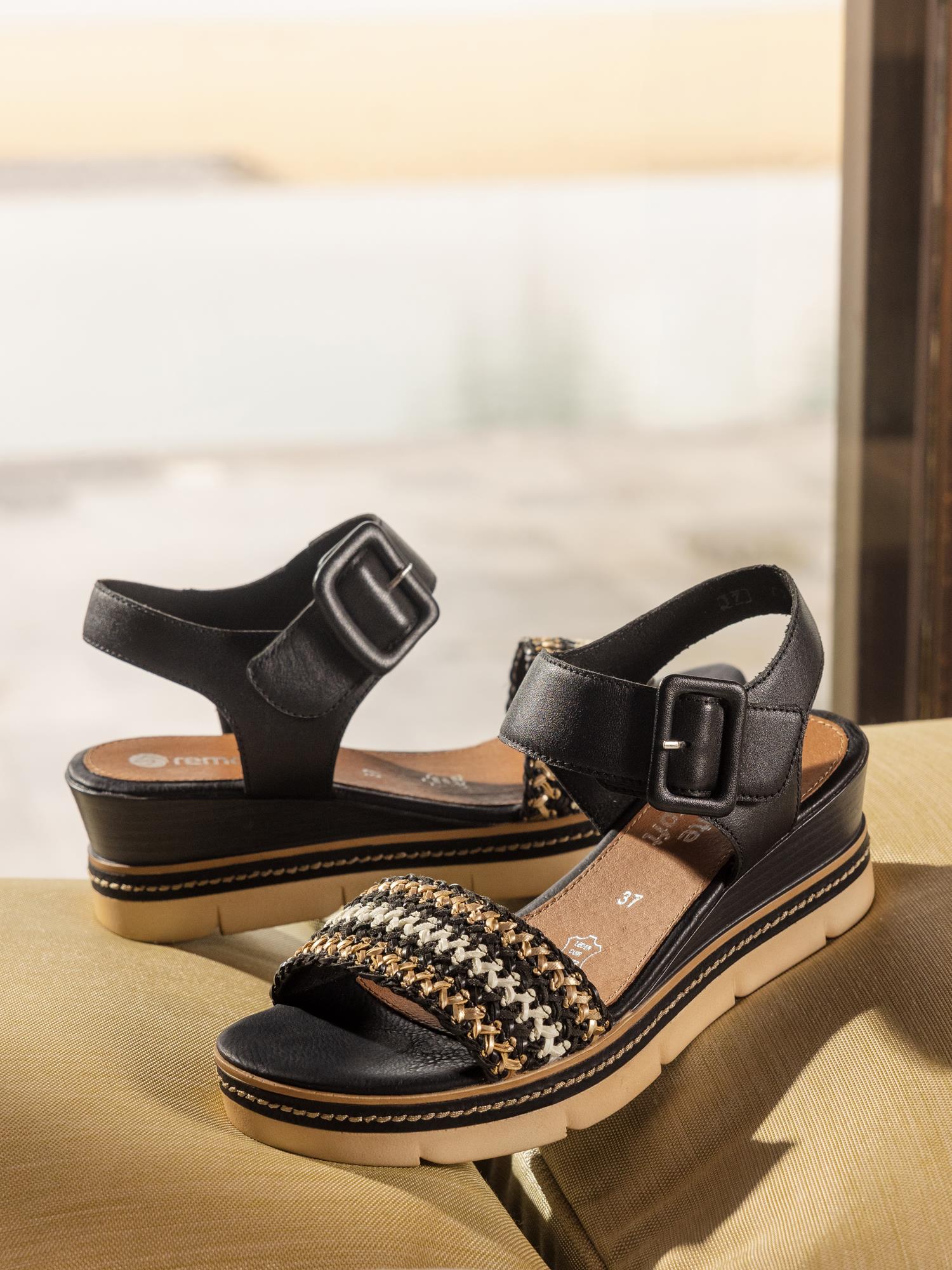 To svarte damesandaler, 03146 Remonte Alvora Sandal fra Remonte, har ankelstropper med store spenner og teksturerte remmer foran. De er vist på en myk overflate, har solbrune såler og er belyst av naturlig lys mot en uskarp bakgrunn.