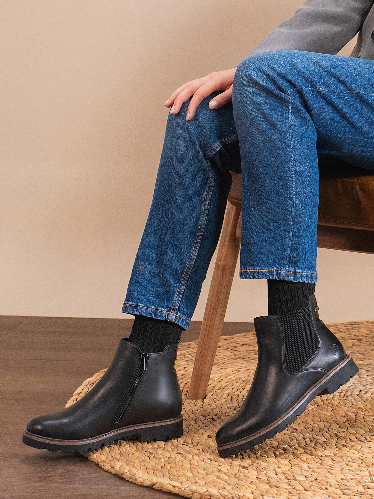 En person sitter på en trestol i blå jeans og svarte sokker, iført Remonte 04571 Torino Leather Ankle Boots, med det ene benet krysset over det andre på et sirkelformet vevd teppe.