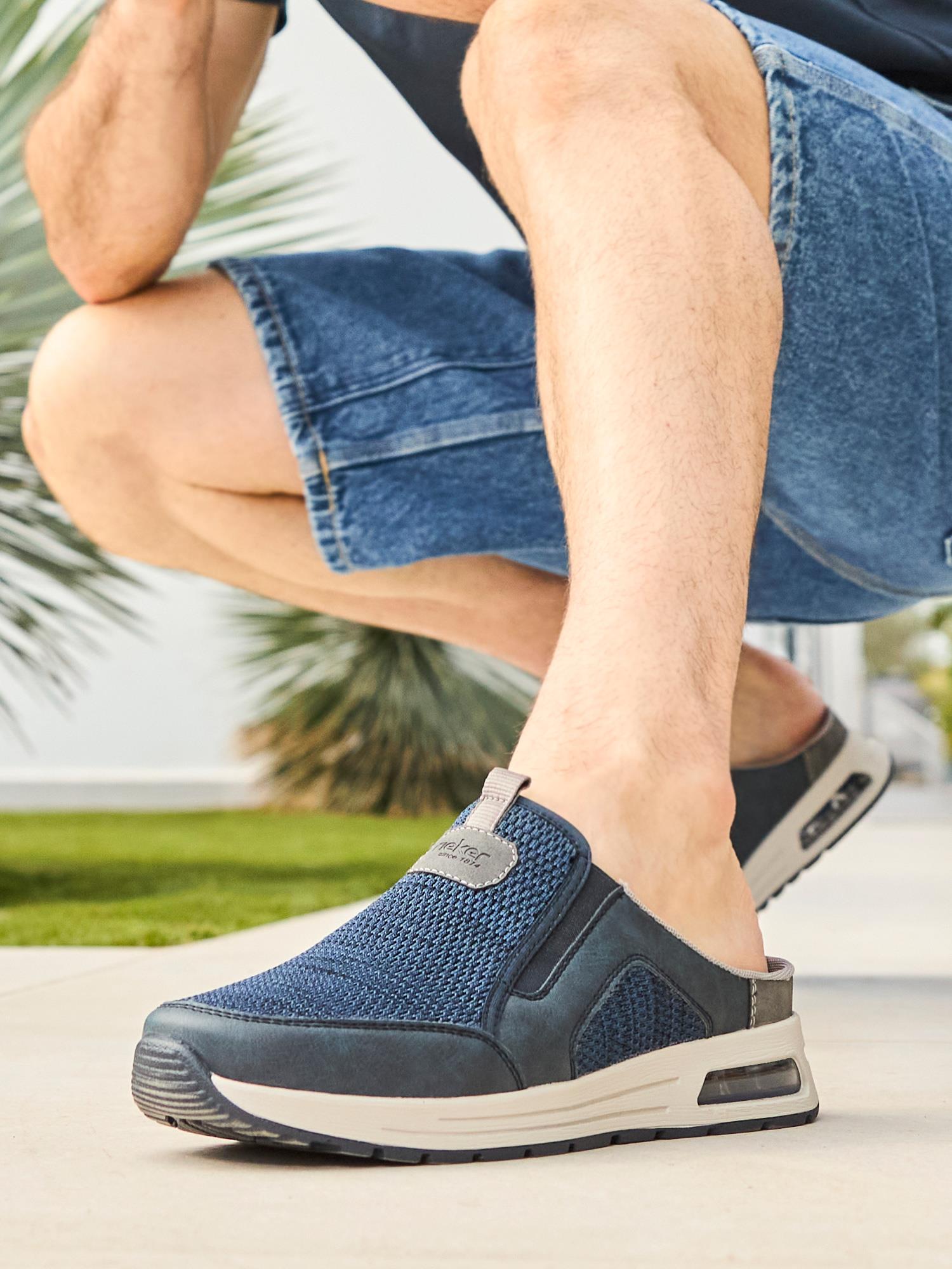 Iført blå 03051 Rieker Nolan Slip-on joggesko fra Rieker Comfort & Style og denimshorts sitter en person på huk utendørs blant grøntområder og moderne landskapsarkitektur, og nyter komforten på en solrik dag.