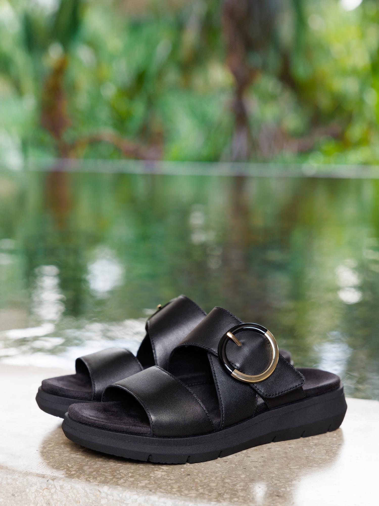 Et par svarte Remonte 03141 Selma-sandaler i vegansk skinn, med brede reimer og en stor gullspenne, ligger på en lys flate ved et basseng med uskarpt grønt løvverk bak.