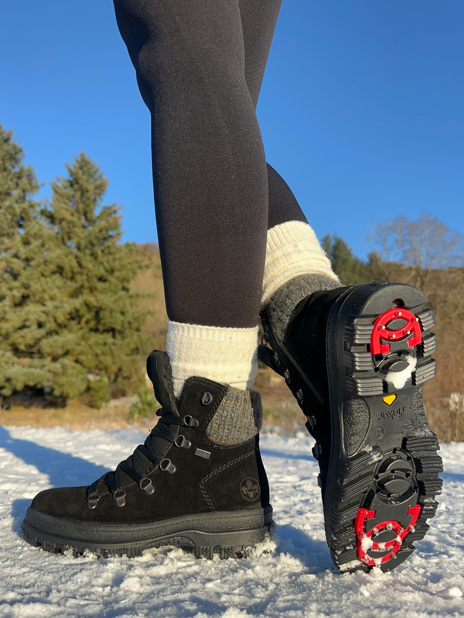 Nærbilde av en person iført 05364 Rieker Varmfòret vintersko med brodder fra Rieker Comfort & Style, svarte leggings og kremfargede sokker, stående på snø med grønne furutrær og blå himmel i bakgrunnen.
