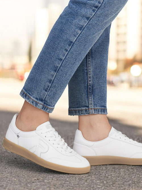 En person iført blå jeans og Rieker Sport 03486 San Diego Leather Sneaker Women White med lysebrune såler står på en asfaltert flate utendørs; bybygninger skimtes i bakgrunnen, noe som understreker Swing2Go-komforten.