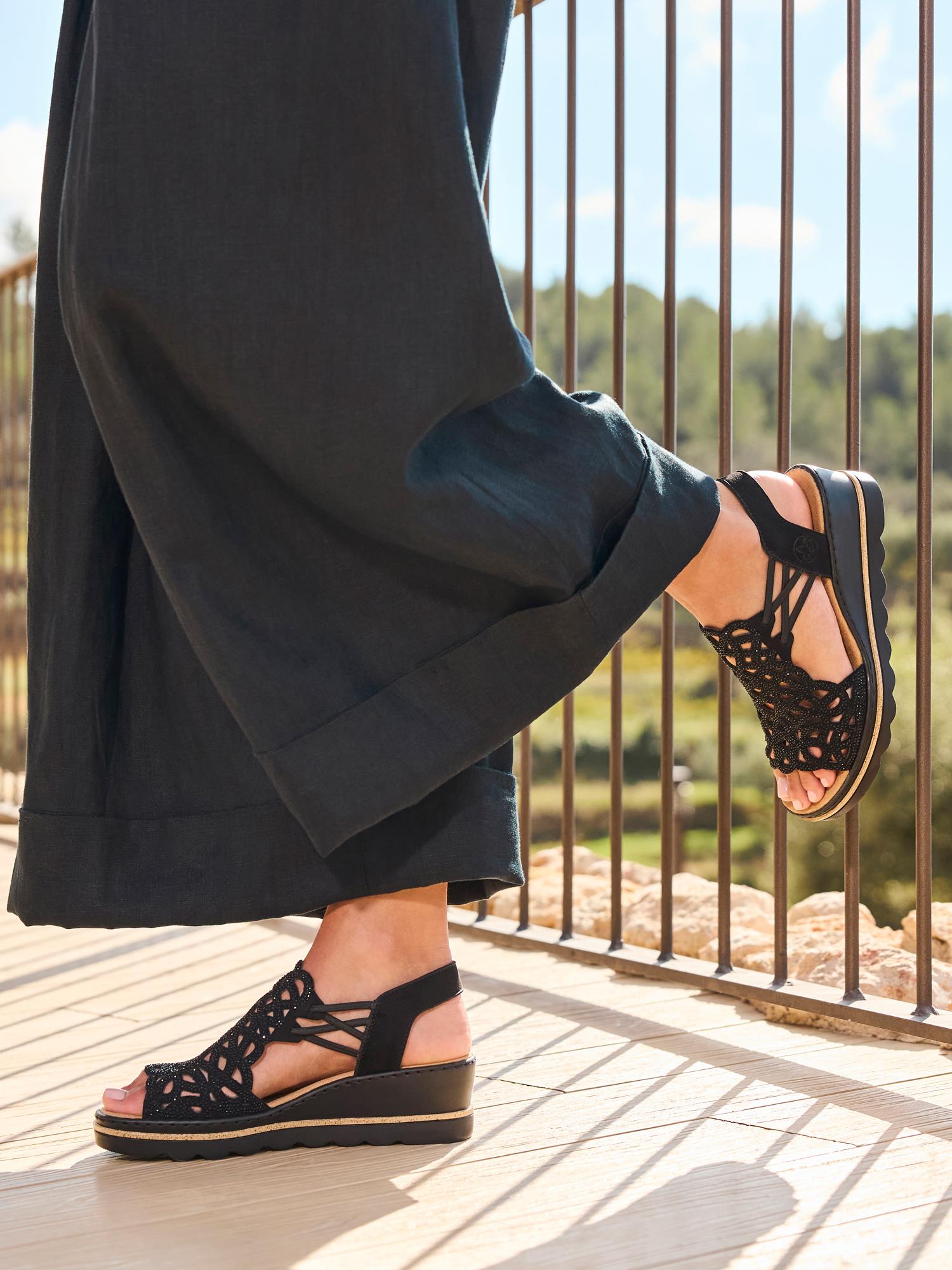 En person i sorte bukser med vide ben og 03003 Rieker Sorella Sandal fra Rieker Comfort & Style står på en solfylt balkong med et metallrekkverk og frodig grønt i bakgrunnen.