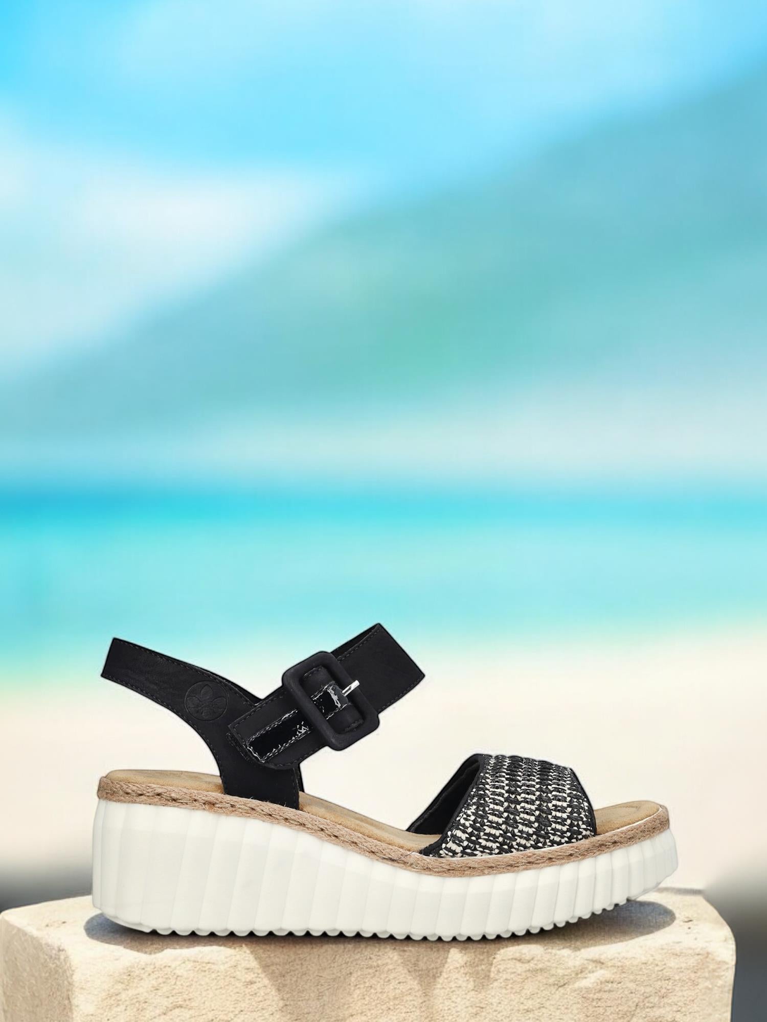 Den svarte 03025 Rieker Celensa Sandal fra Rieker Comfort & Style, med en vevd kile, tykk hvit såle, spennerem og komfortbredde G for ekstra komfort, vises på en stein med en uskarp strand og hav i bakgrunnen.