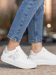 En person i lyseblå jeans har på seg hvite Rieker Sport 03461 ComfortFlex Leather joggesko med sølvfarget P-logo og antistress-teknologi, mens han står på en asfaltert flate mot en uskarp bybakgrunn.