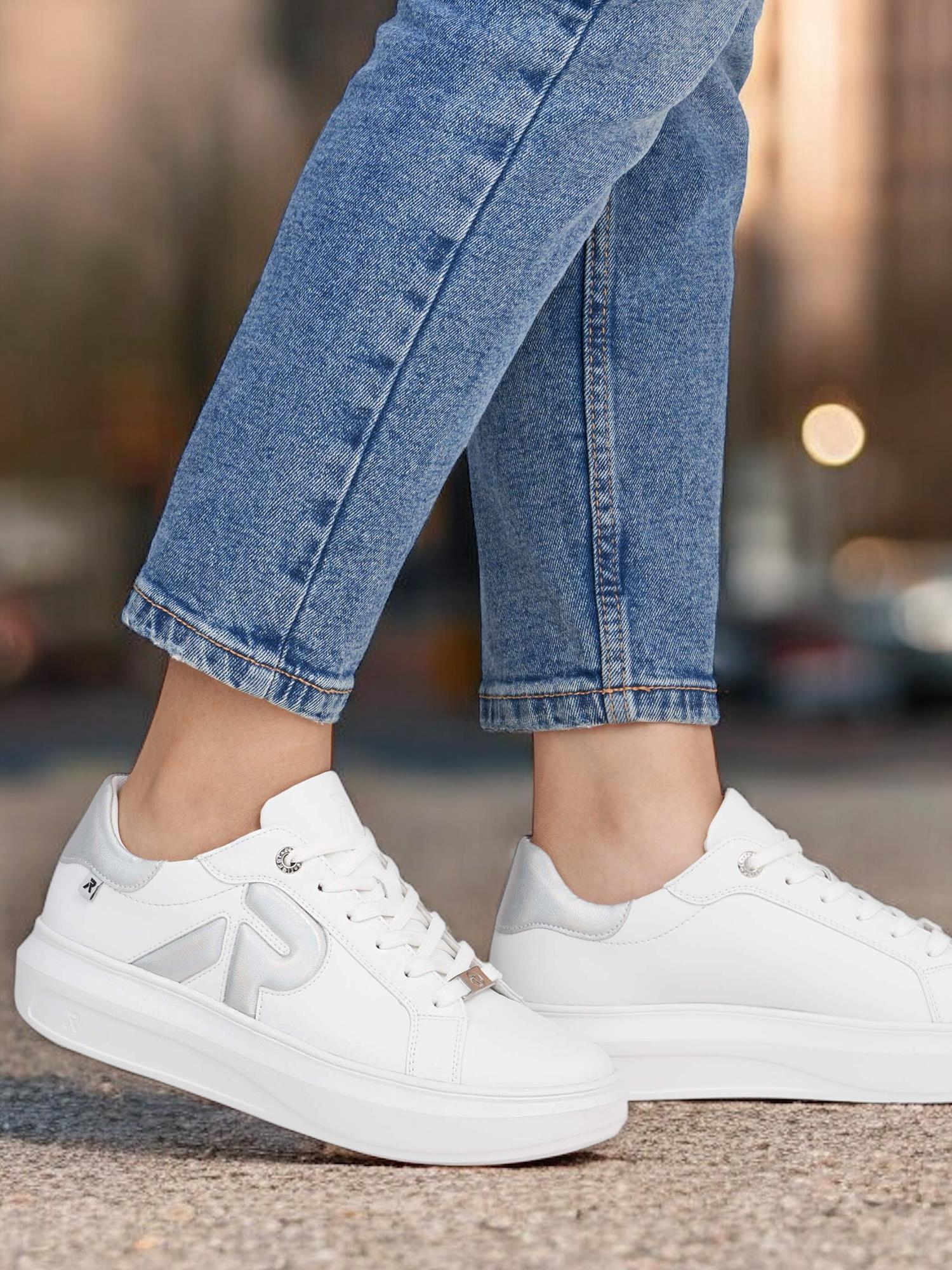 En person i lyseblå jeans har på seg hvite Rieker Sport 03461 ComfortFlex Leather joggesko med sølvfarget P-logo og antistress-teknologi, mens han står på en asfaltert flate mot en uskarp bybakgrunn.