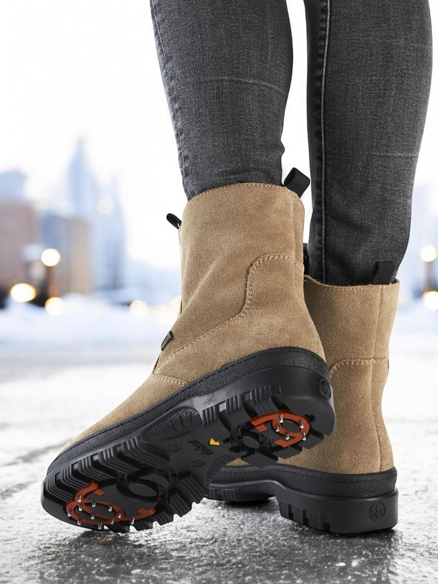 Nærbilde av en person iført 05039 Rieker Leather Tex Varmfôret med brodder fra Rieker Comfort & Style, som viser solbrune støvler med svart såle, oransje grepdetaljer og vanntett membran, stående i en snødekt bygate.