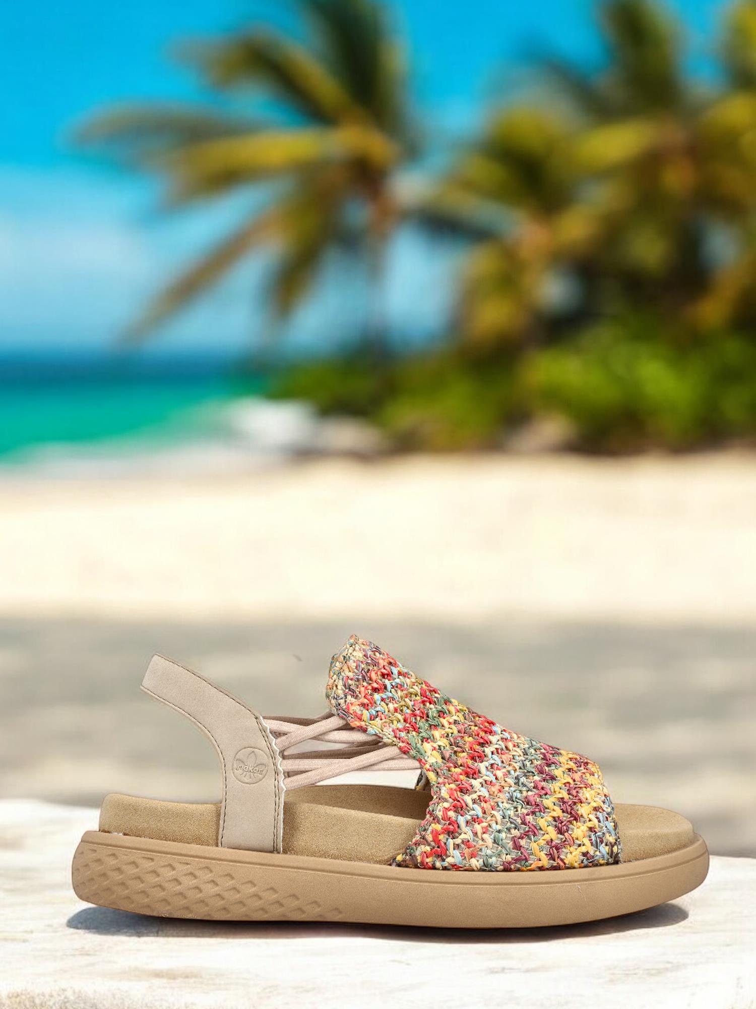 03341 Rieker Viala Sandal fra Rieker Comfort & Style, med et fargerikt komfortdesign og beige såle, vises på hvitt med en uskarp tropisk strandbakgrunn, noe som fremhever den ultralette konstruksjonen.